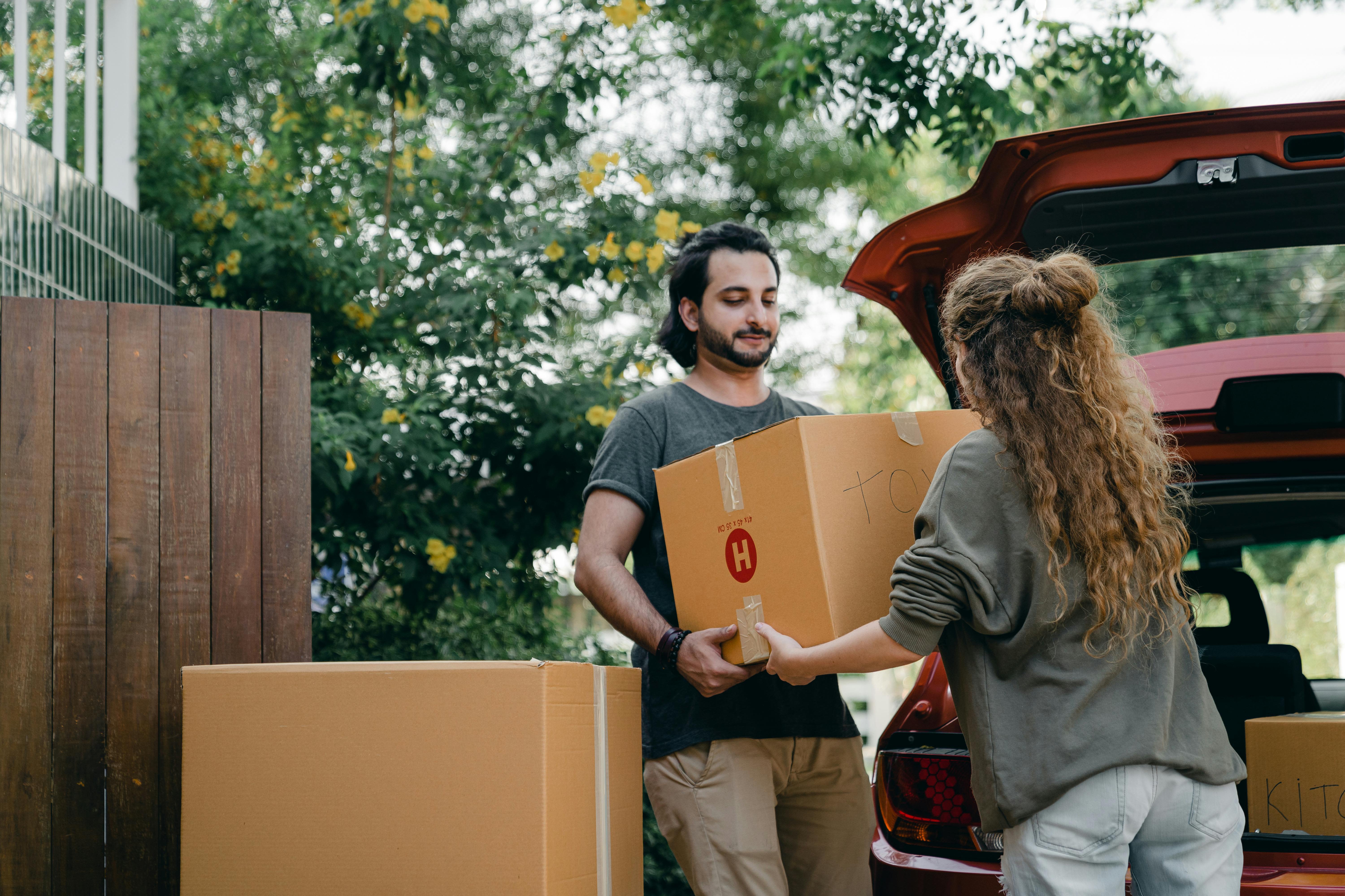 Two people loading essential moving day survival kit and first night boxes into the back of a red car, preparing to keep important items within easy reach.
