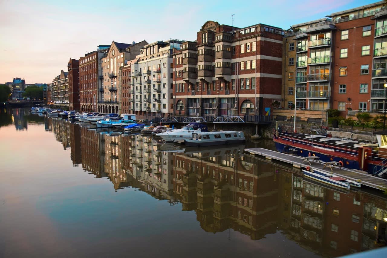 View of Bristol harbour at sunset, featuring historic and modern buildings reflected in the calm water, with moored boats and houseboats lining the quay.