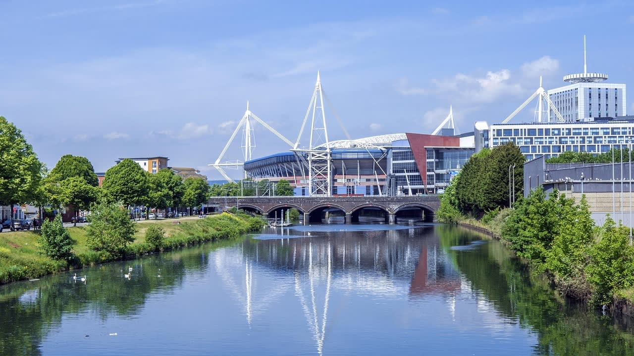 View of the Principality Stadium and city buildings along the River Taff in Cardiff, with a bridge crossing the river and trees lining the banks on a sunny day.