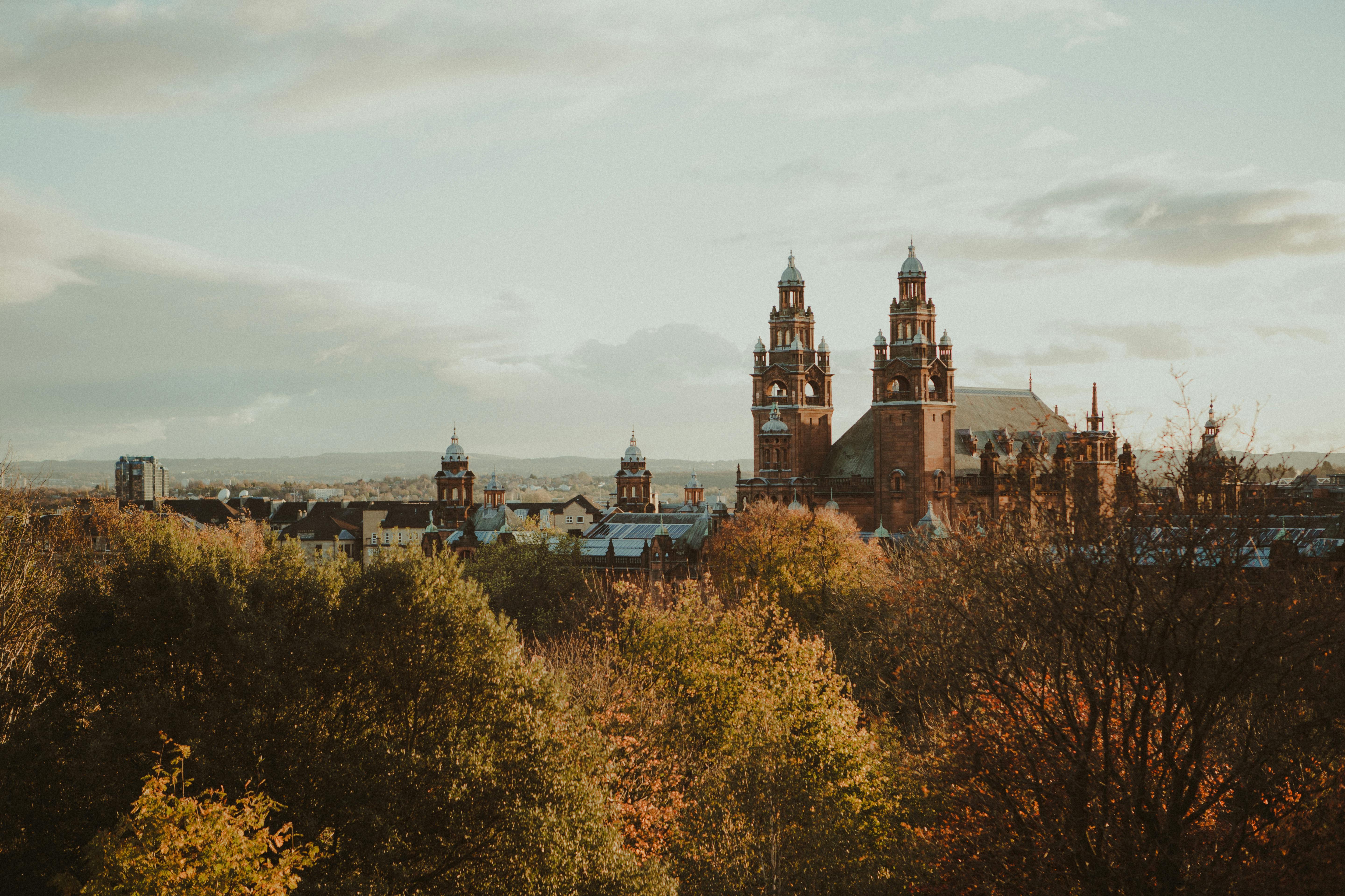 Panoramic view of Glasgow at sunset, featuring the iconic Kelvingrove Art Gallery and Museum with its distinctive red sandstone towers, surrounded by autumn trees and the city skyline stretching into the distance.