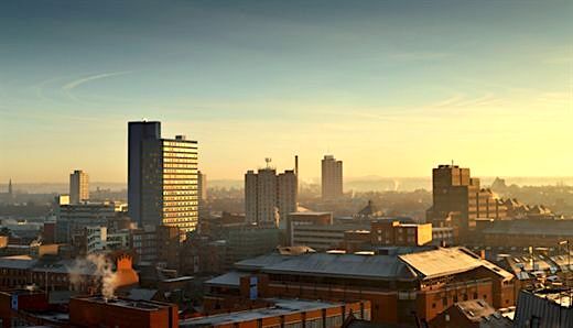 Skyline of Leicester city at sunrise, featuring a mix of modern high-rise buildings and older brick structures under a clear sky with a warm golden glow.