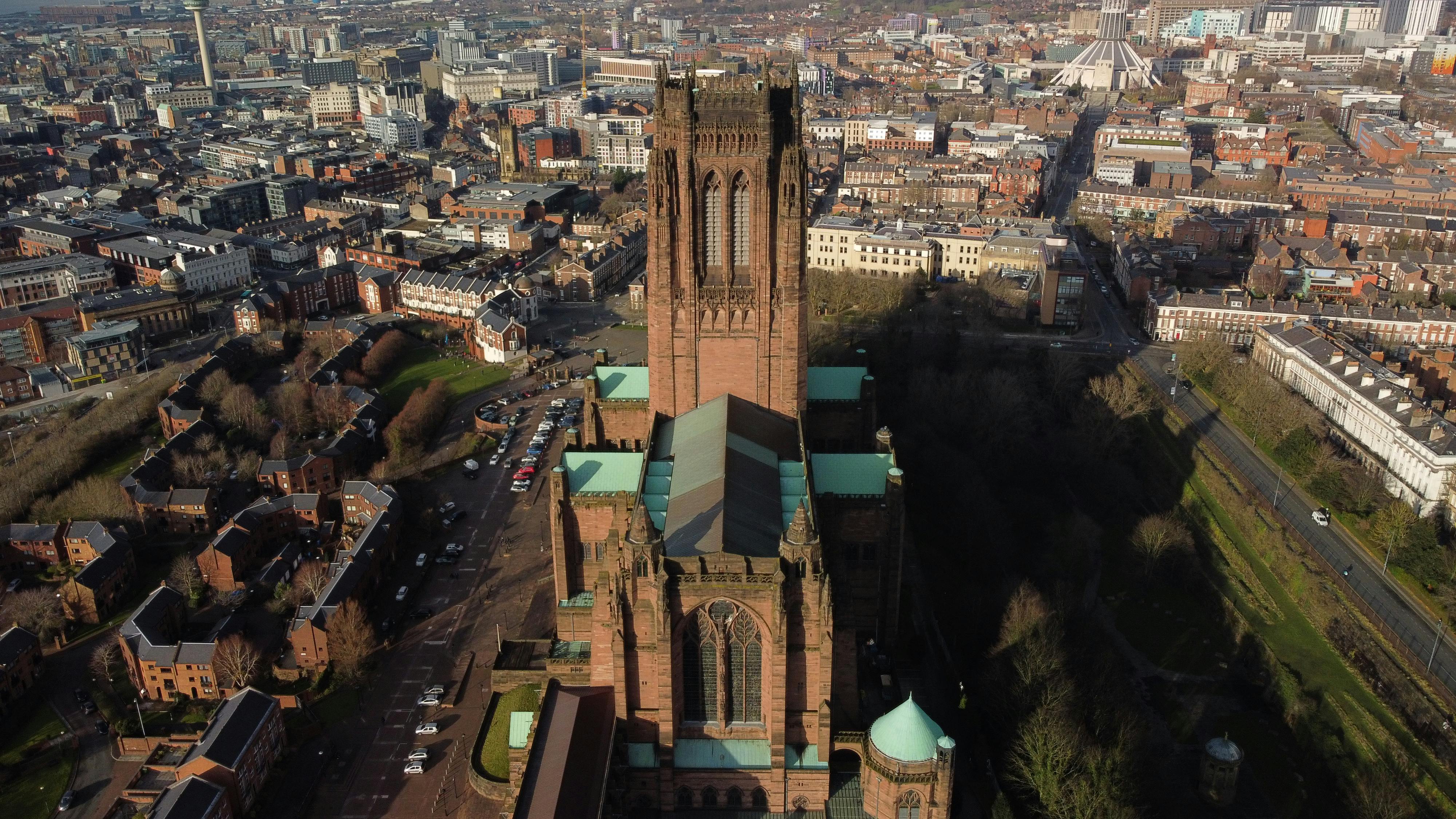 Aerial view of Liverpool Cathedral, a large sandstone structure with greenish copper roofs, standing prominently in the foreground. The cathedral is surrounded by residential streets, green spaces, and a dense urban cityscape. Liverpool landmarks such as 