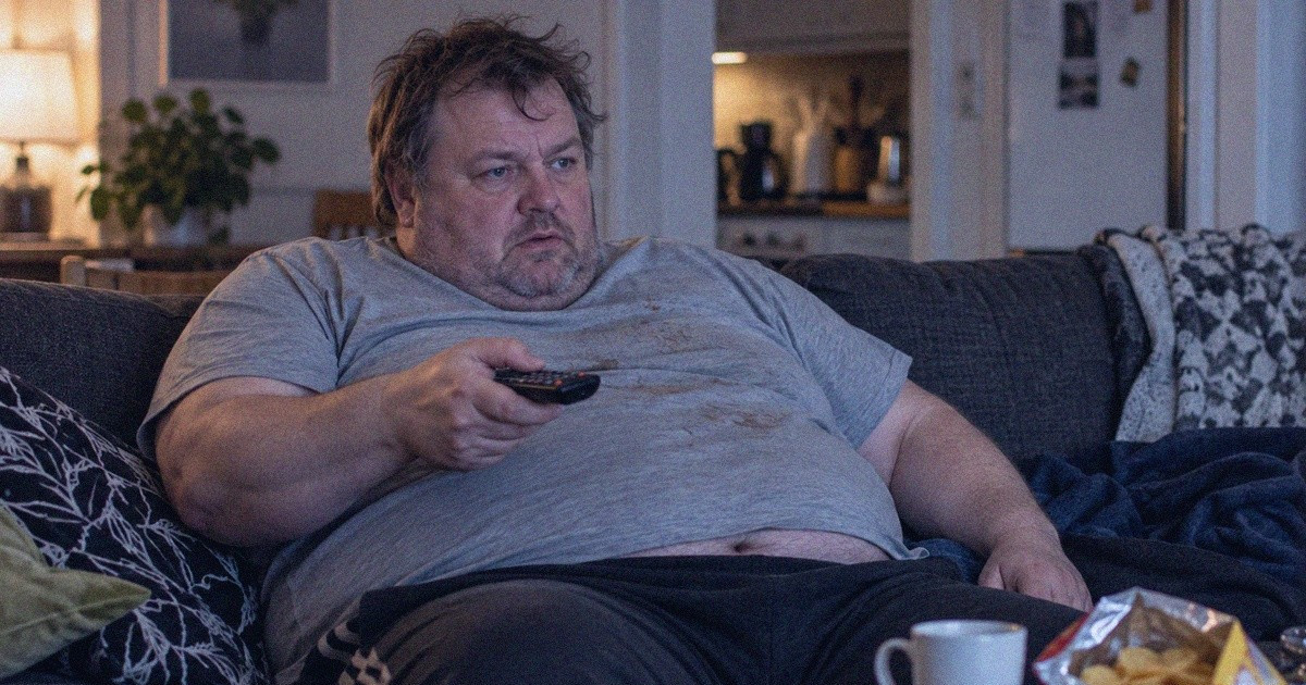 Mies istuu sohvalla kaukosäädin kädessään ja katsoo televisiota.