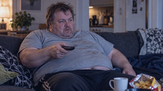 Mies istuu sohvalla kaukosäädin kädessään ja katsoo televisiota.