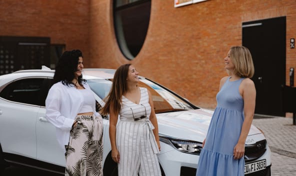 Drei Frauen stehen lachend vor einem Auto.