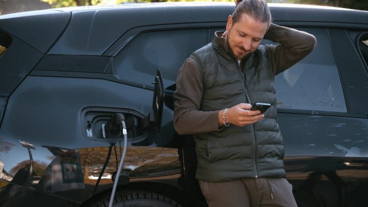Mann lädt E-Auto und schaut aufs Handy.