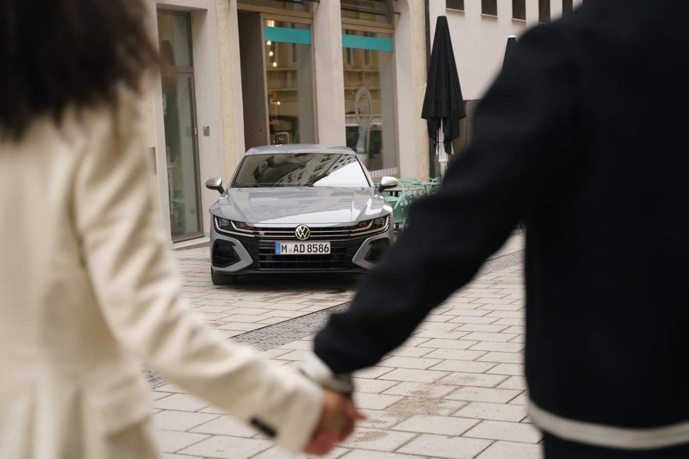 Ein Mann und eine Frau gehen Händchen halten auf einen VW Arteon zu.