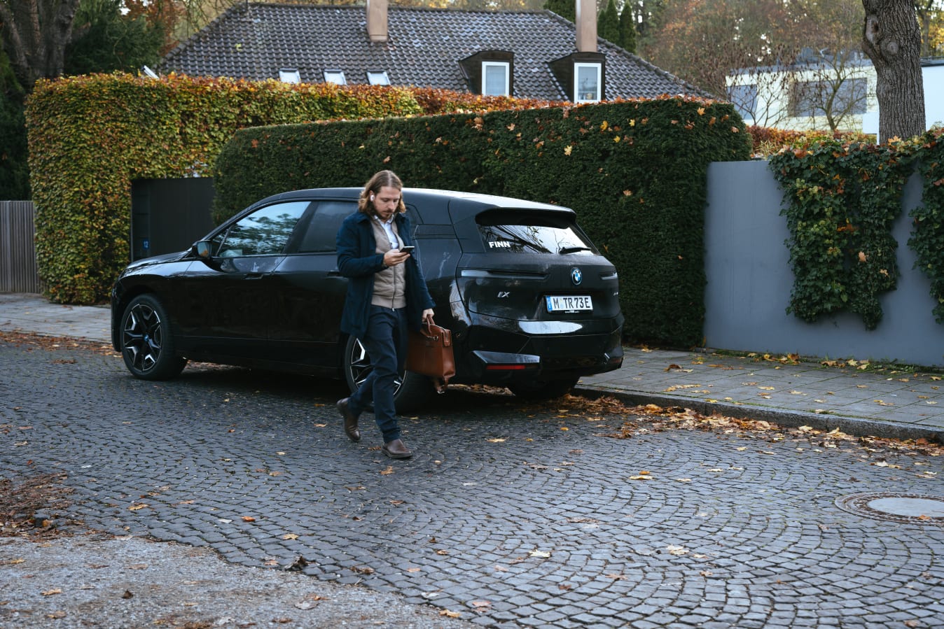 Ein Mann mit Aktentasche schaut auf sein Handy und geht vor seinem FINN Auto.