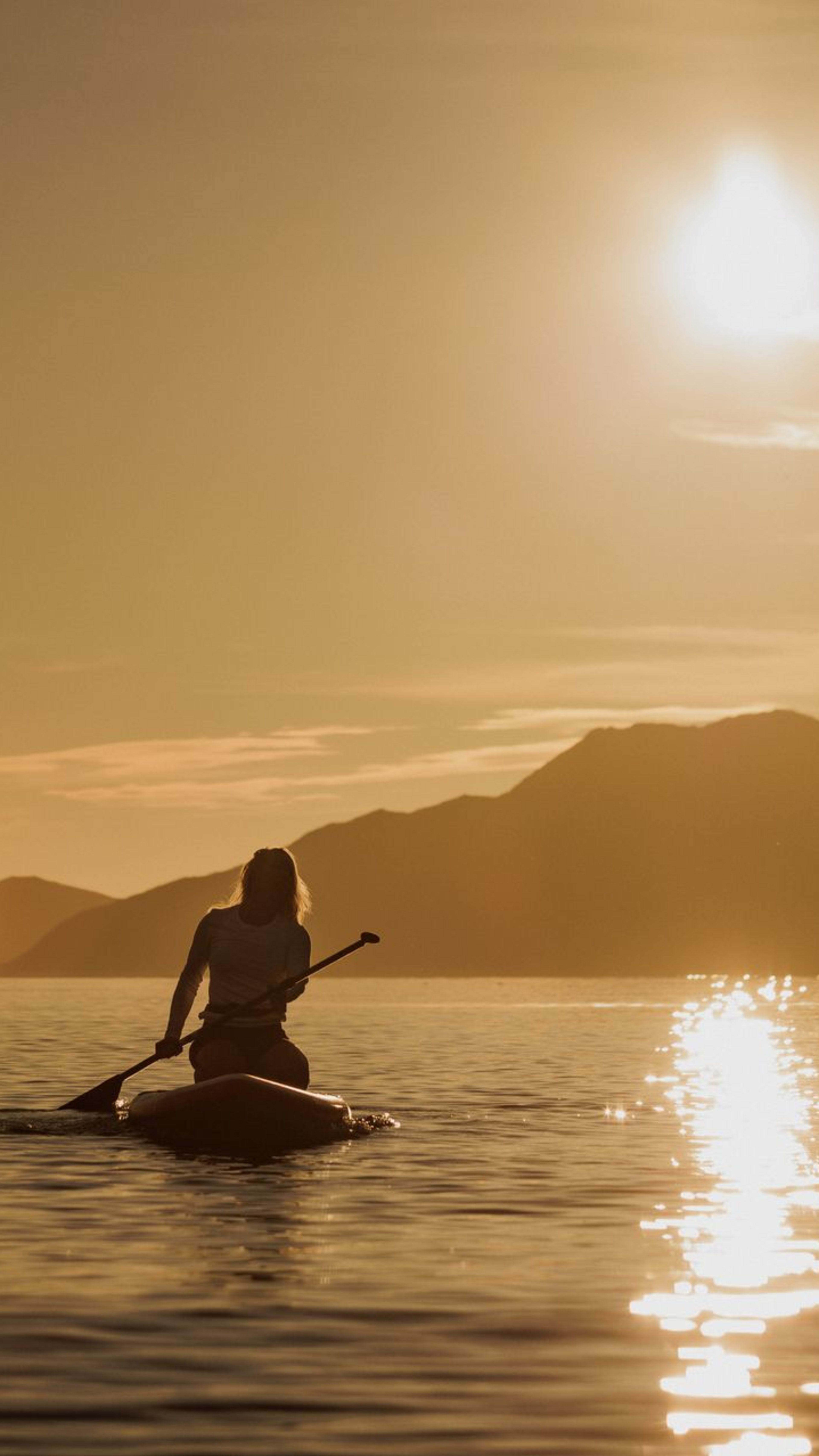 Frau beim Kajakfahren nahe der Insel Kvaløya in Tromsø während der Mitternachtssonne, mit goldenem Sonnenlicht, das sich in ruhigem arktischem Gewässer spiegelt.