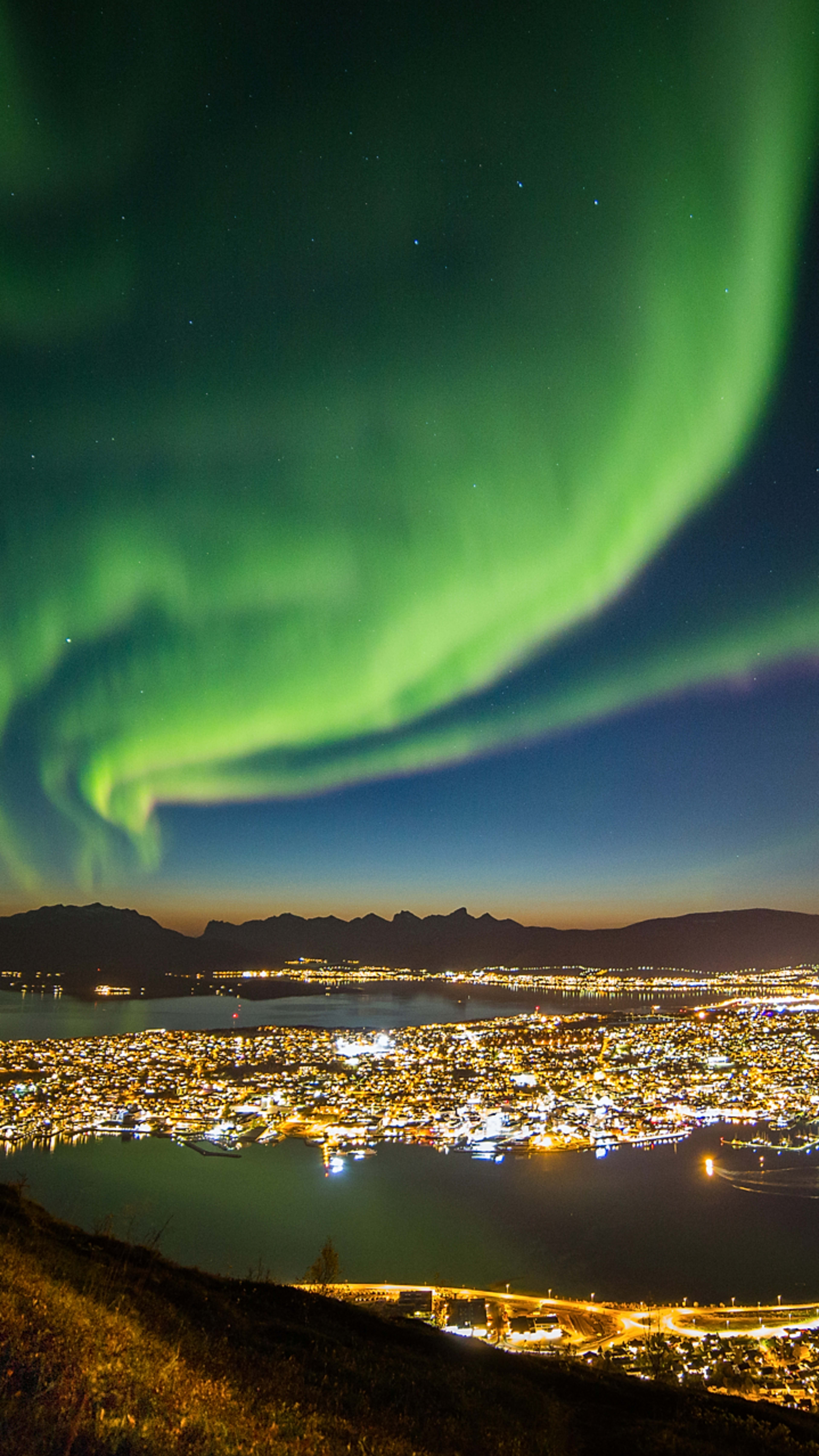 Grüne Nordlichter tanzen am Herbsthimmel über Tromsø, mit Blick auf die beleuchtete Stadt, Fjorde und Berge.