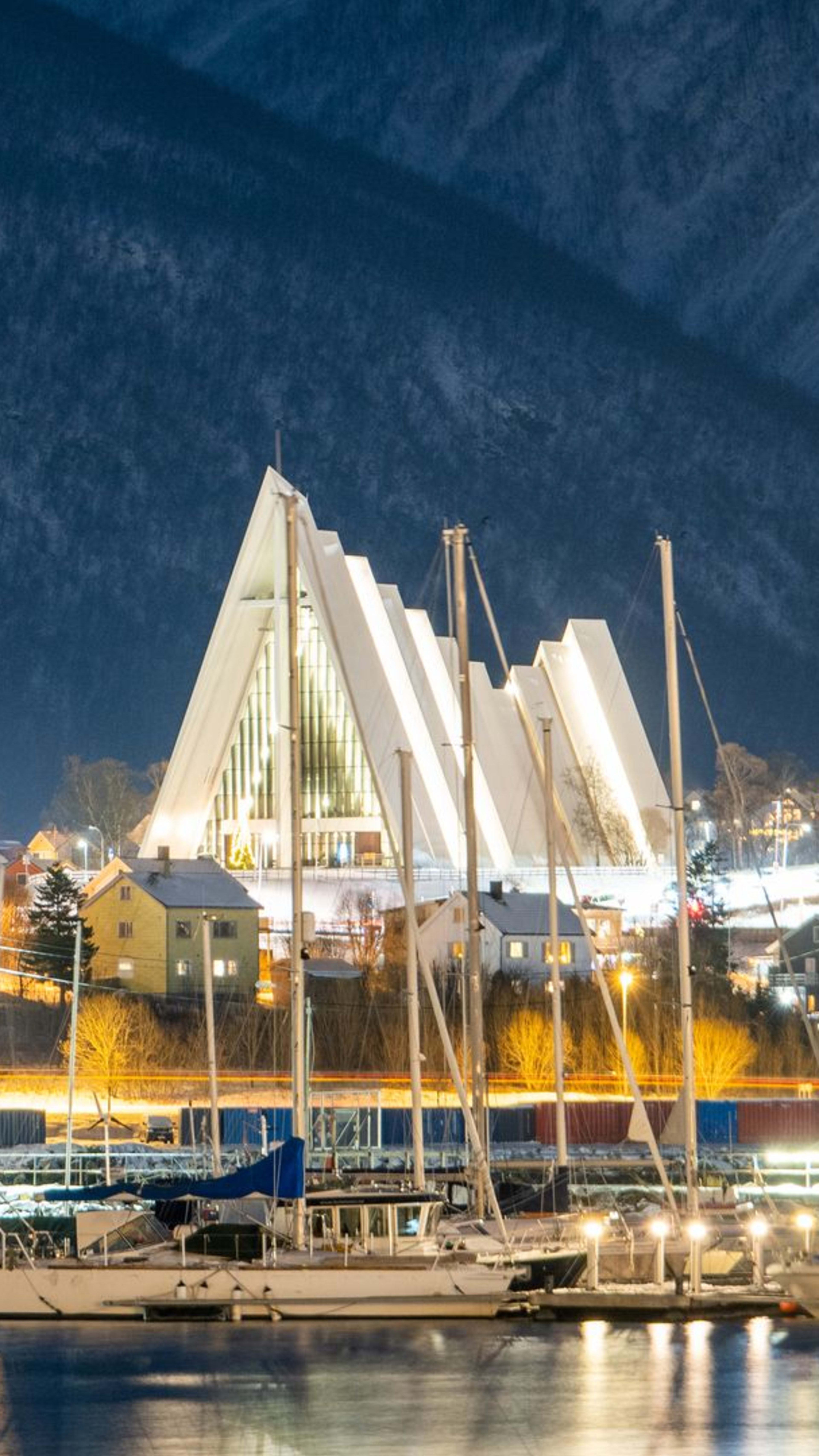 Die beleuchtete Eismeerkathedrale in Tromsø bei Nacht, mit Fischerbooten im Hafen darunter und schneebedeckten Bergen, die sich im Hintergrund erheben.