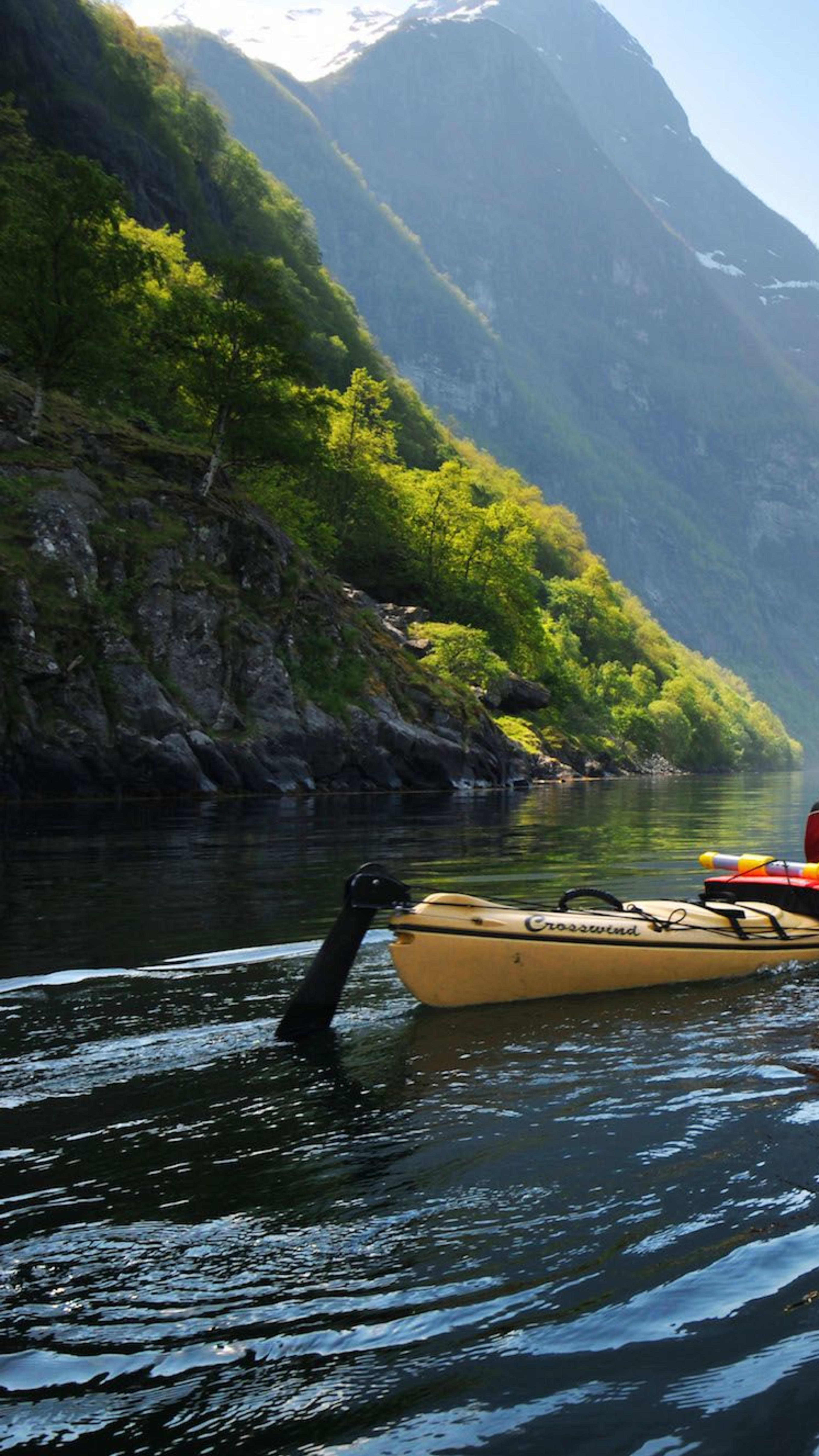 Zwei Kajakfahrer paddeln entlang eines ruhigen, spiegelnden Fjords, umgeben von hohen grünen Bergen und dramatischen Klippen unter einem klaren blauen Himmel.