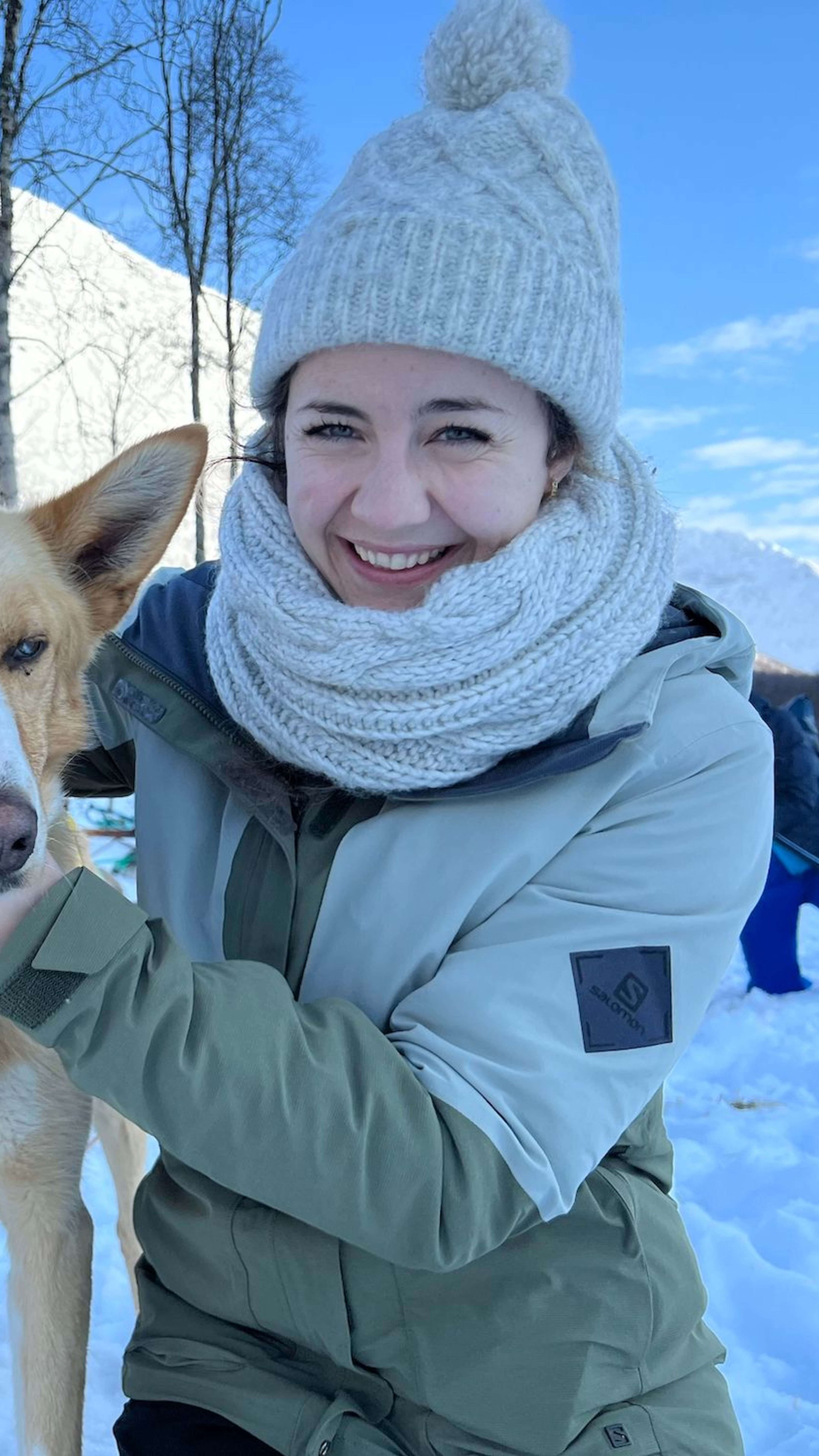 Eine Person in warmer Kleidung streichelt zwei Schlittenhunde im Schnee, wobei ein Husky aufmerksam steht und ein brauner Hund ruhig steht.