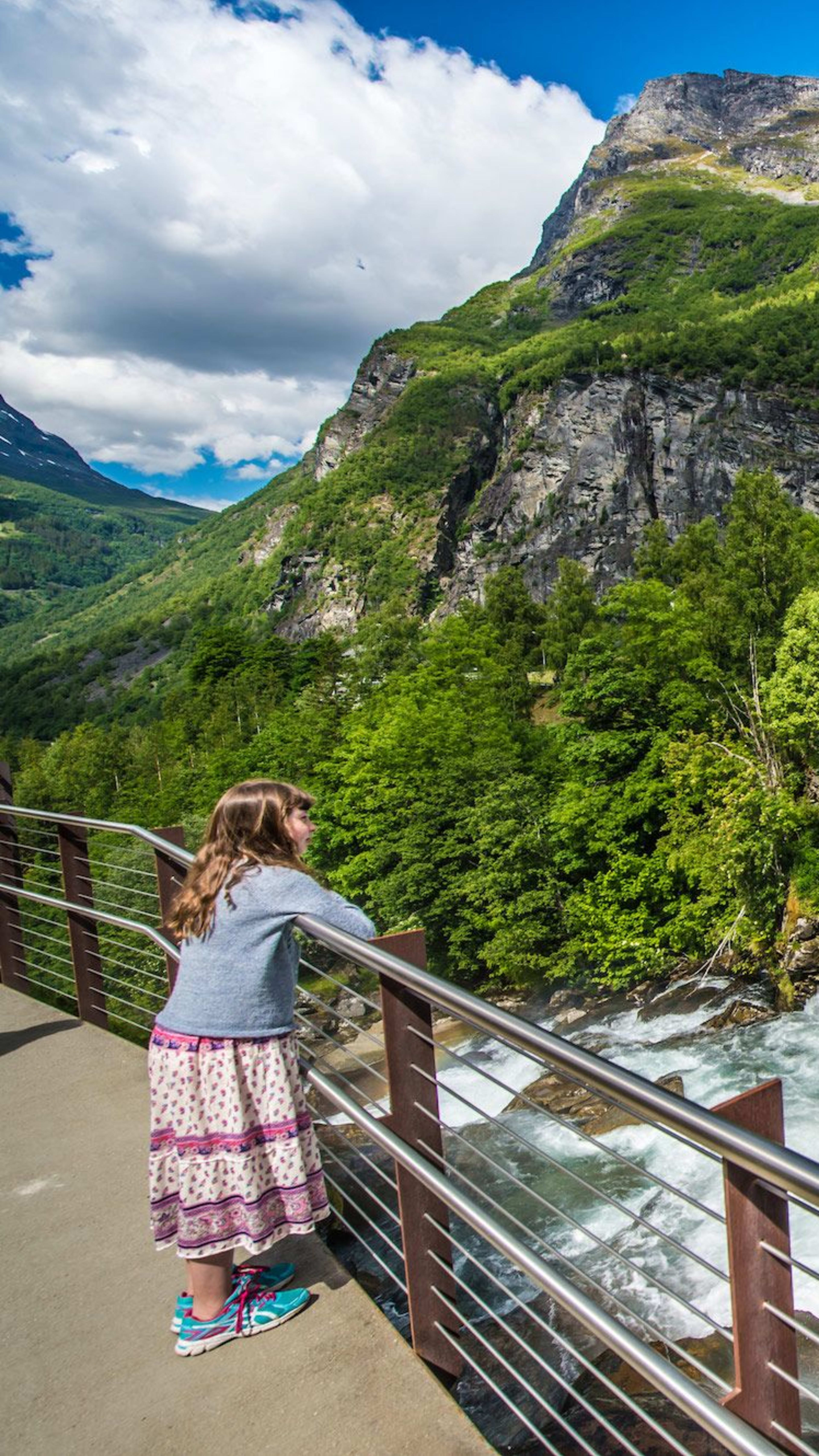 Mädchen lehnt am Geländer und schaut auf Wasserfall in grüner Landschaft unter blauem Himmel.