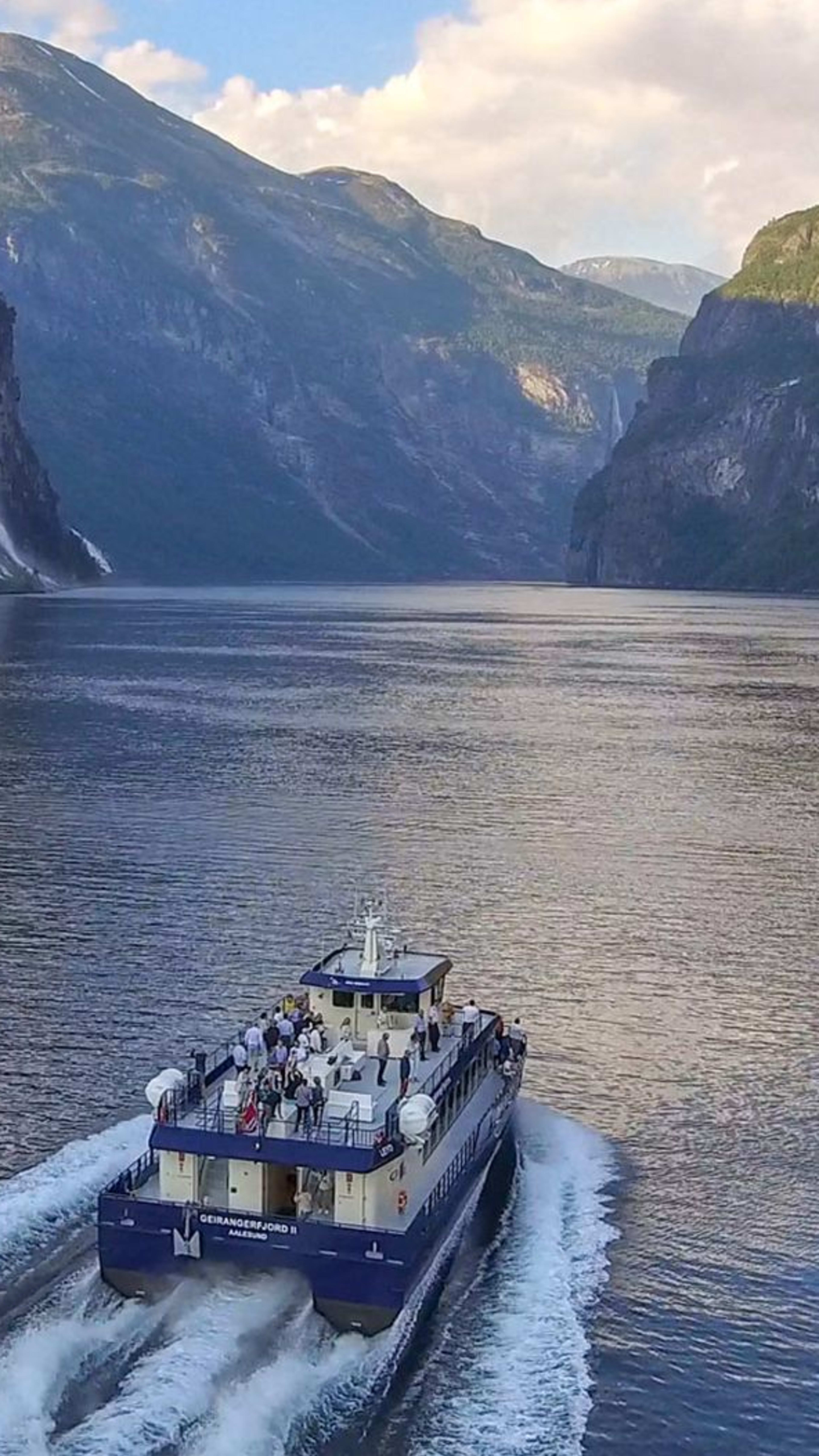 Ausflugsboot im Geirangerfjord mit steilen Klippen, Wasserfällen und grünen Bergen.