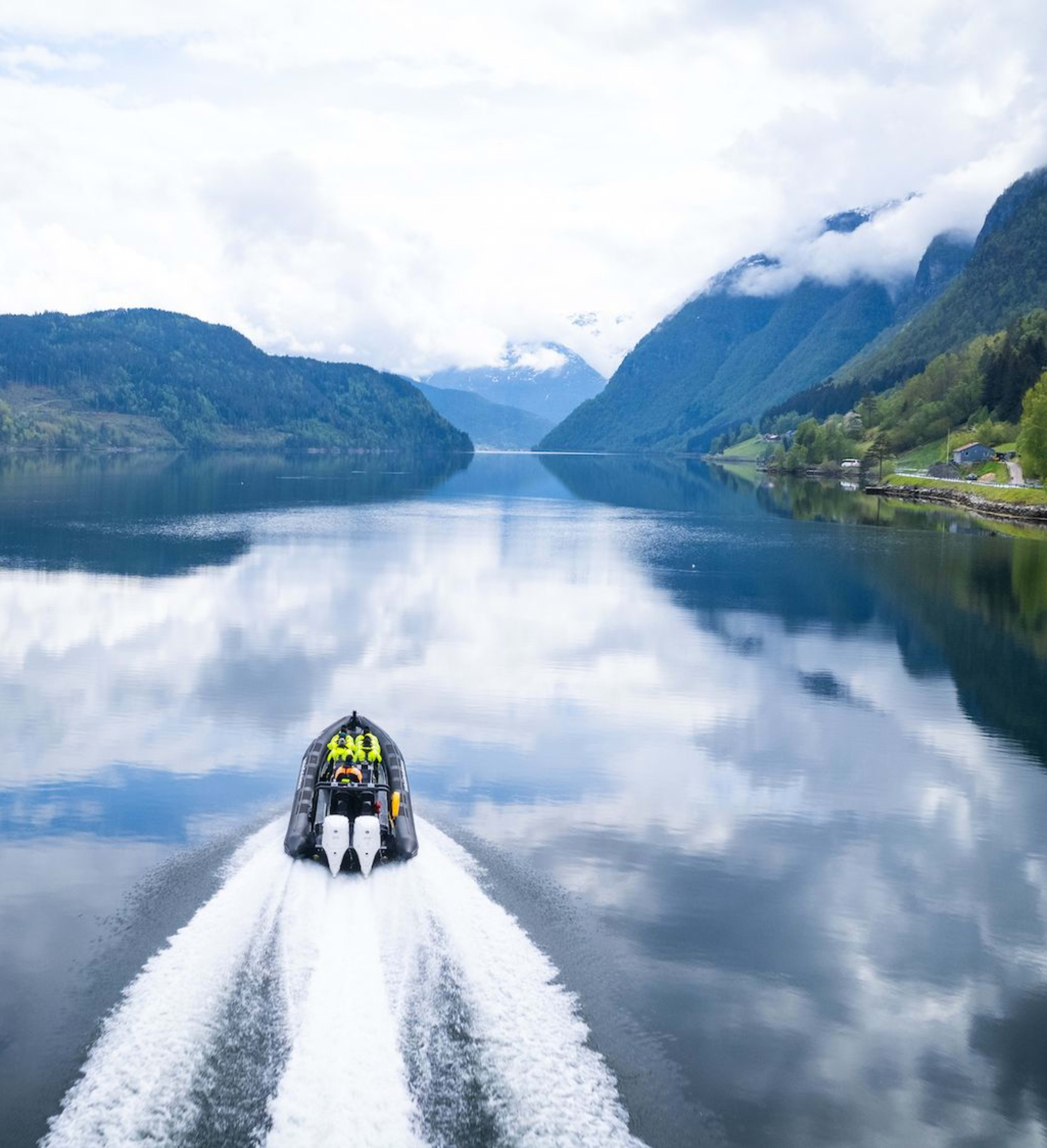 Båt med passasjerer suser over speilblank Hardangerfjord som reflekterer fjell og skyet himmel.