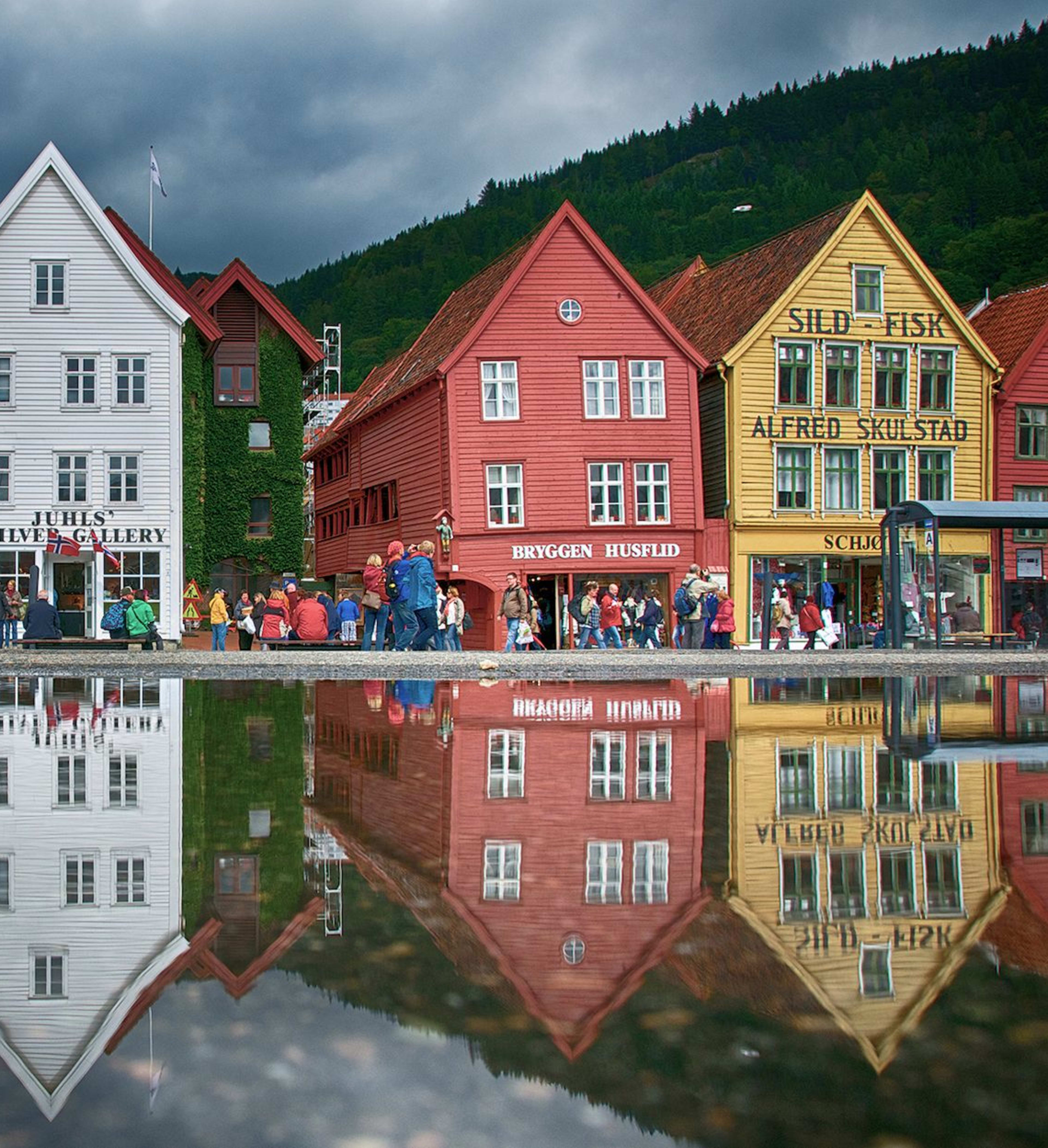 Fargerike Bryggen-bygninger i Bergen speiles i en hav, folk går under dramatiske skyer.