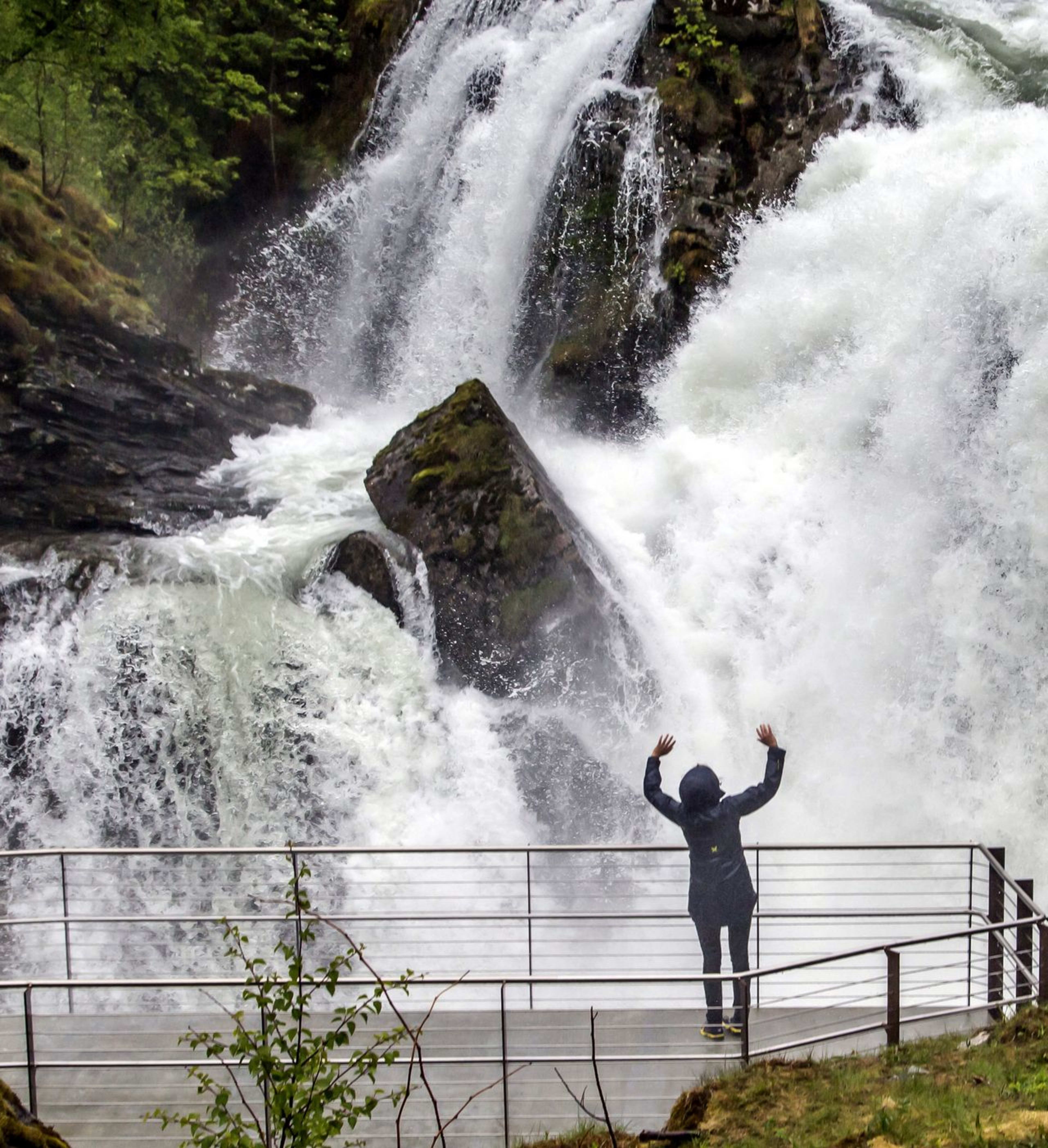Person in rain jacket raises arms facing massive waterfall among greenery.