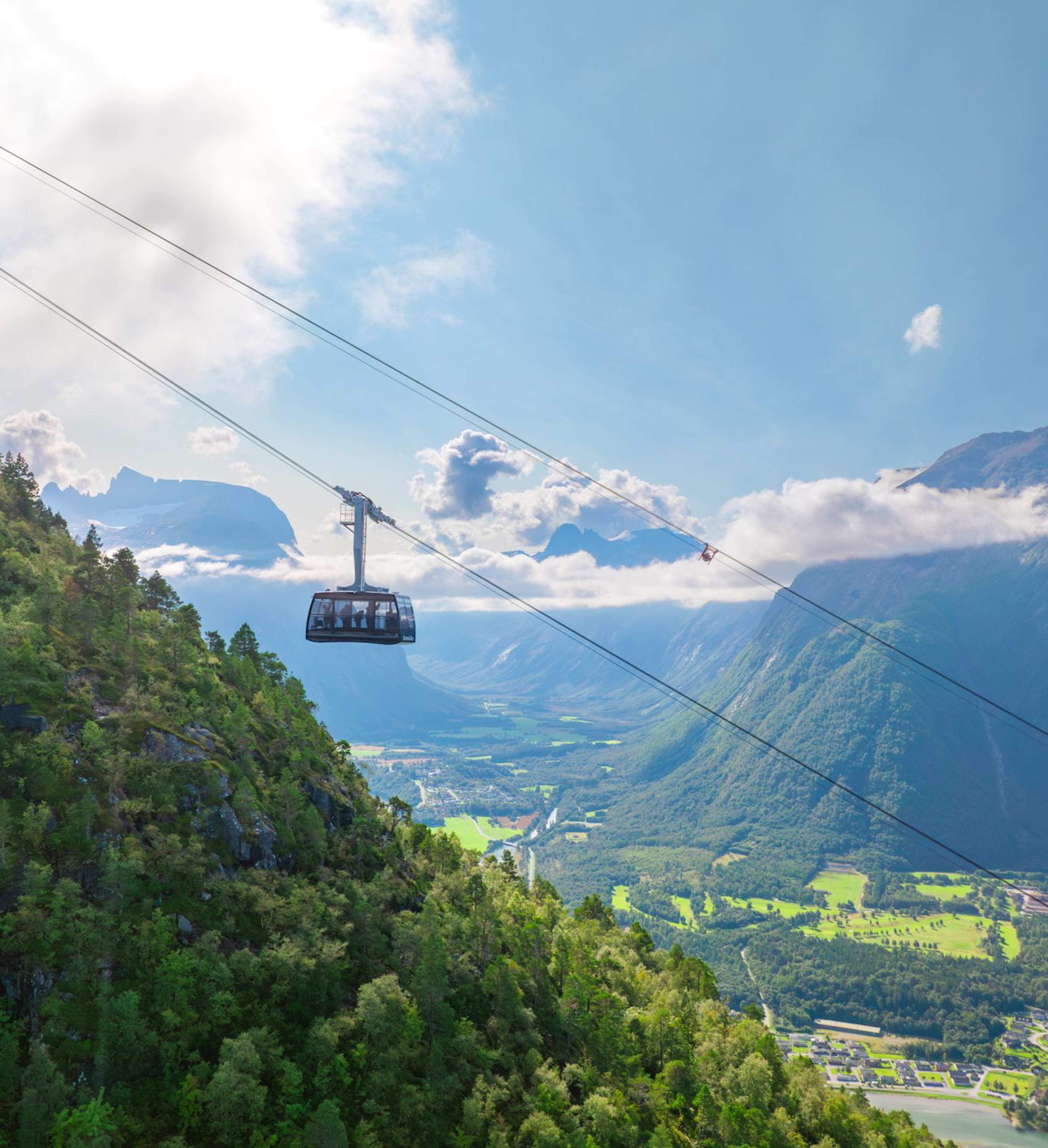 A cable car travels above a lush green mountain, with fields, villages, and winding roads below, and towering mountains in the distance.