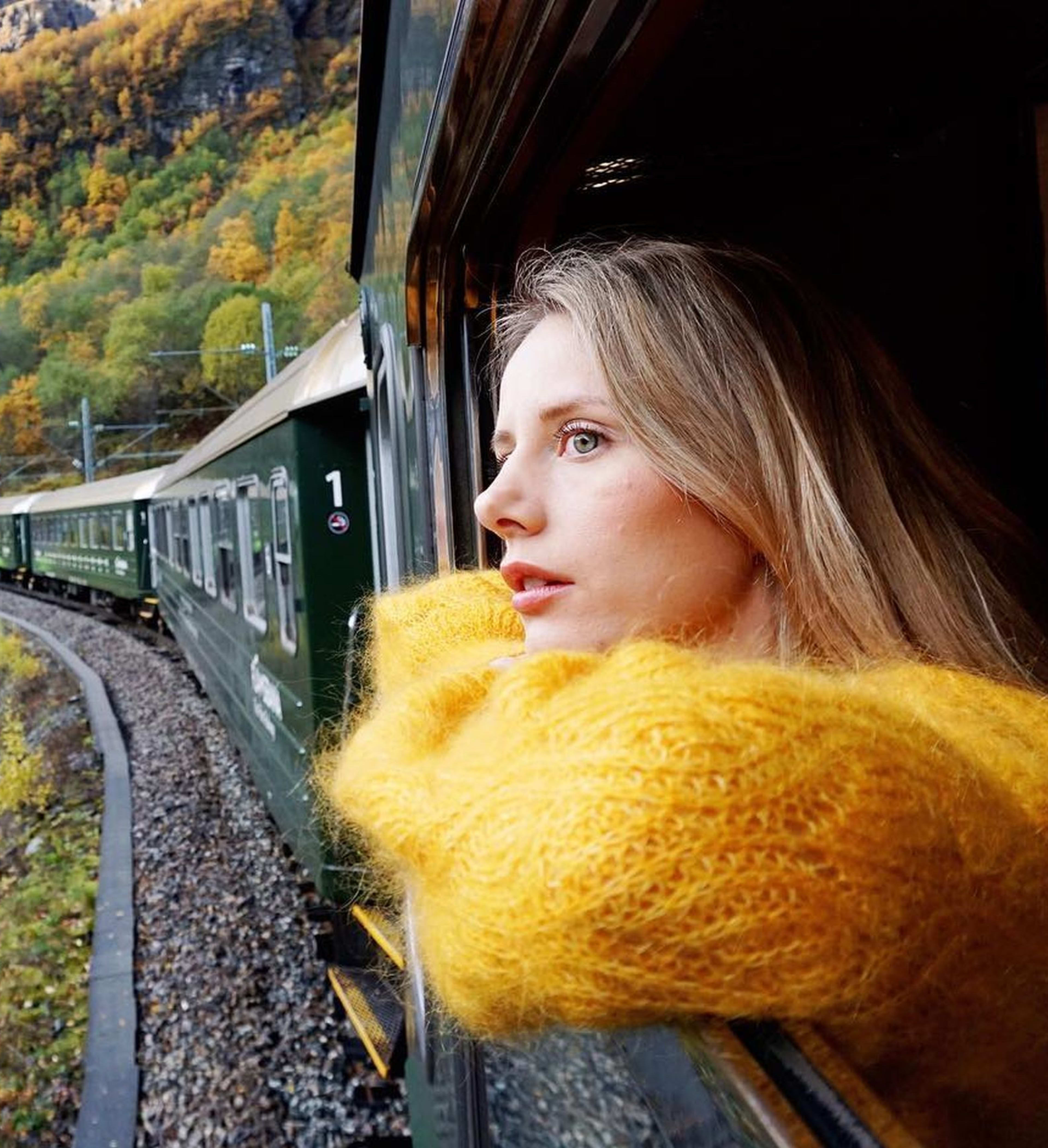 Frau im gelben Pullover blickt aus grünem Zugfenster auf herbstliche Landschaft.