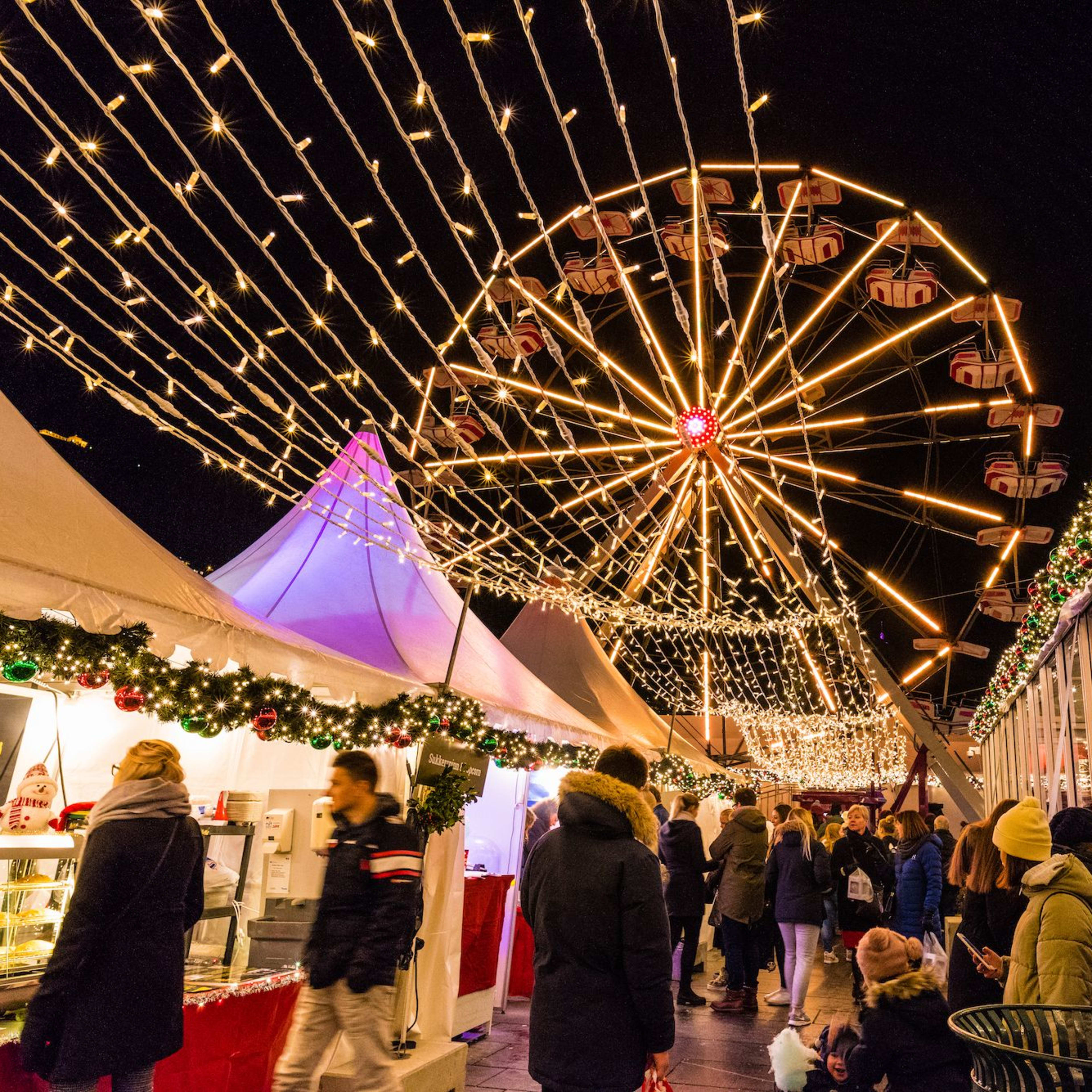Ein belebter Weihnachtsmarkt bei Nacht mit einem leuchtenden Riesenrad und Lichterketten, dekorierten Ständen und Essensständen.