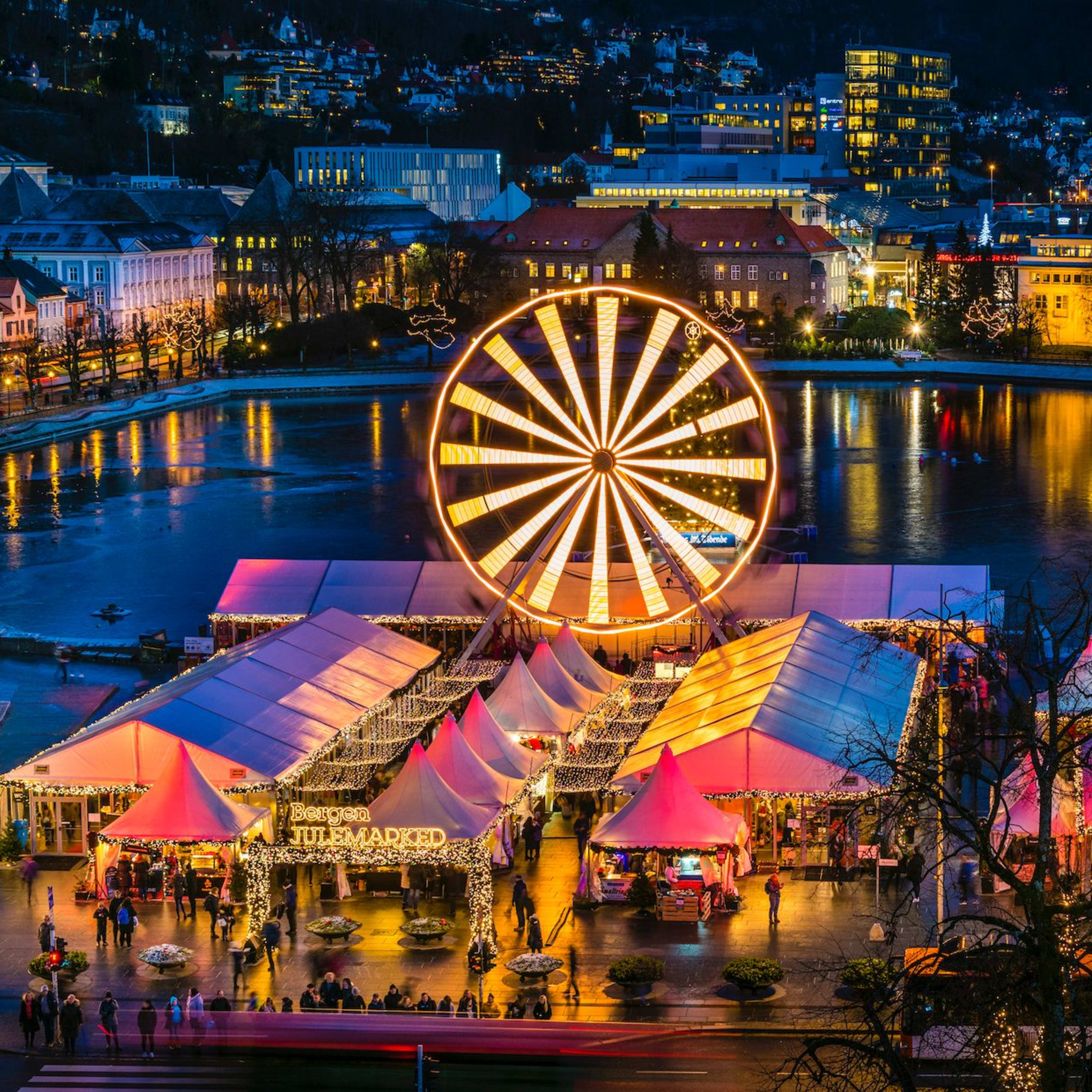 Ein Weihnachtsmarkt bei Nacht mit einem hell erleuchteten Riesenrad, festlichen Zelten und einem ruhigen Gewässer.