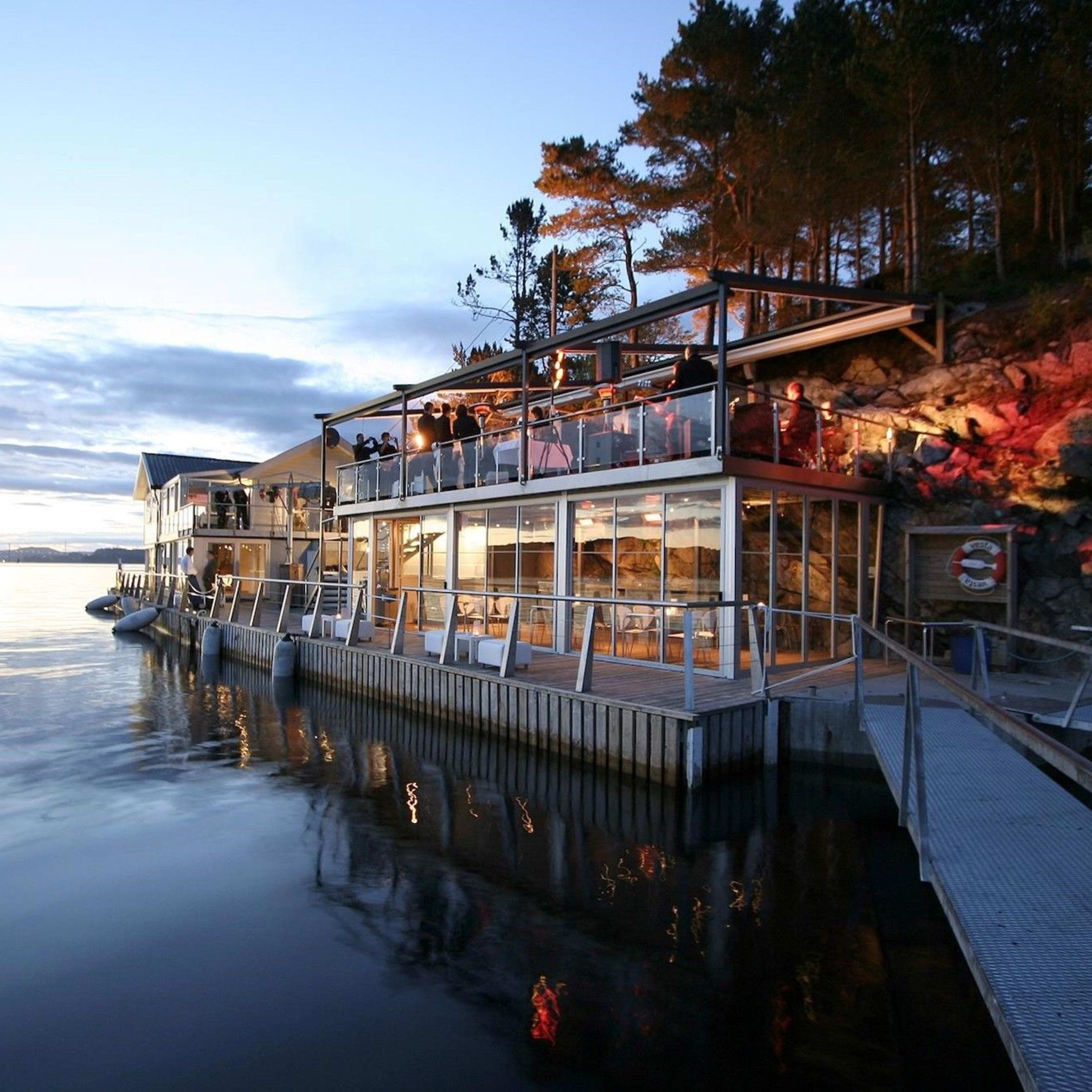 Koselig restaurant med glassfasade ved vannet, varmt lys i skumringen og speilinger på stille vannflate.