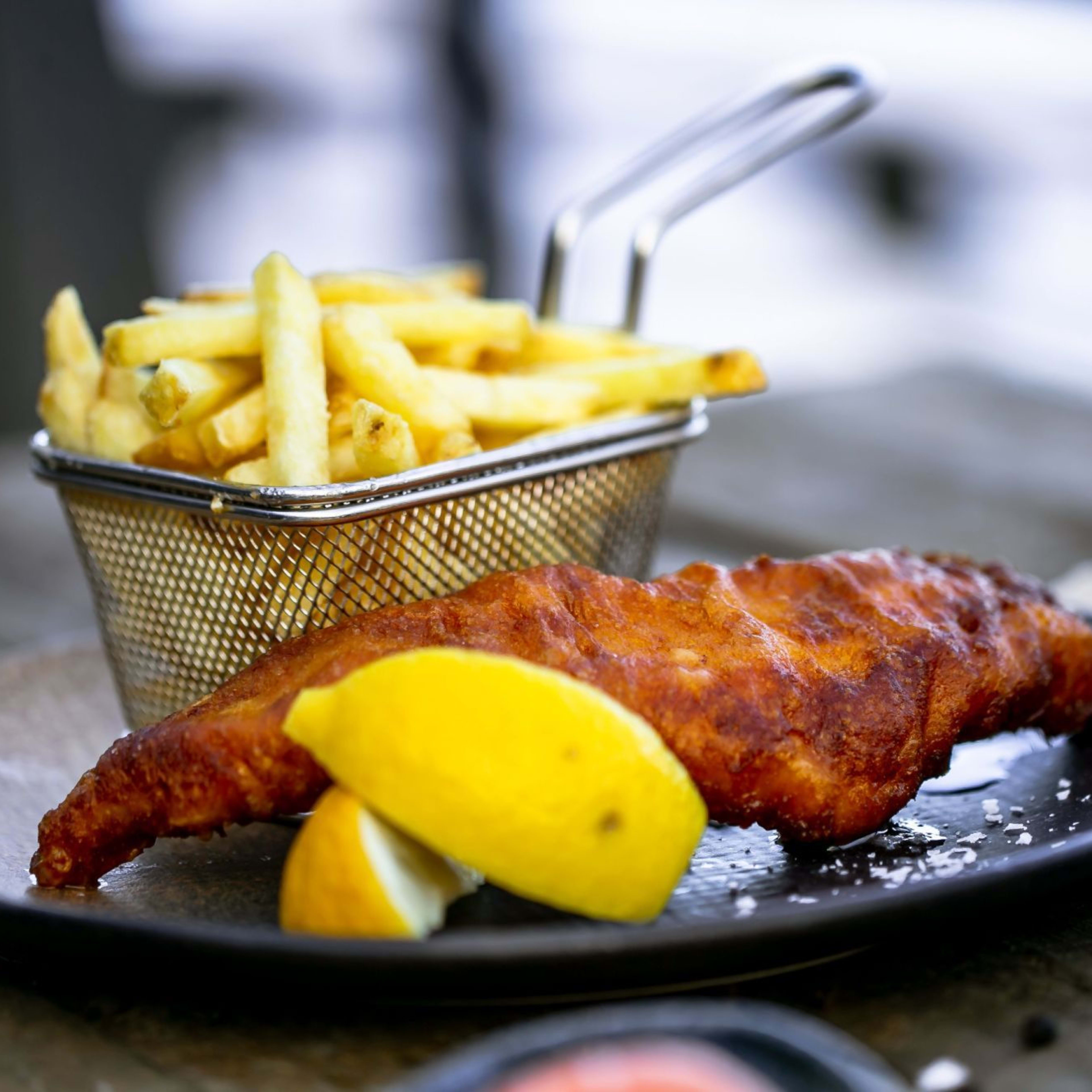 Ein klassisches Gericht Fish and Chips auf einem schwarzen Keramikteller mit Zitronenspalten, Pommes frites und Meersalz.