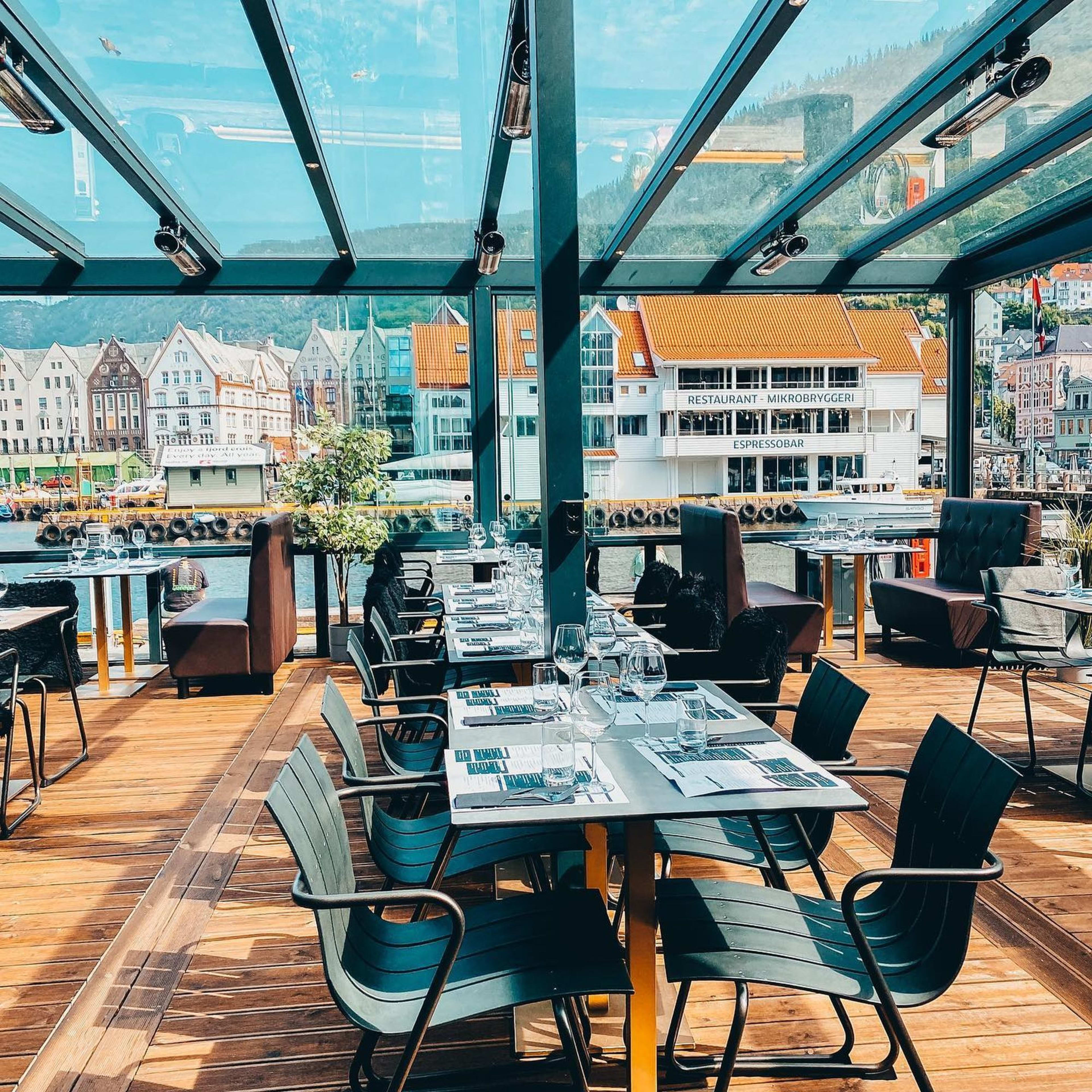 Eine stilvolle Restaurantterrasse mit modernen schwarzen Stühlen und Panoramablick auf die Wasserfront von Bergen und historische Gebäude.