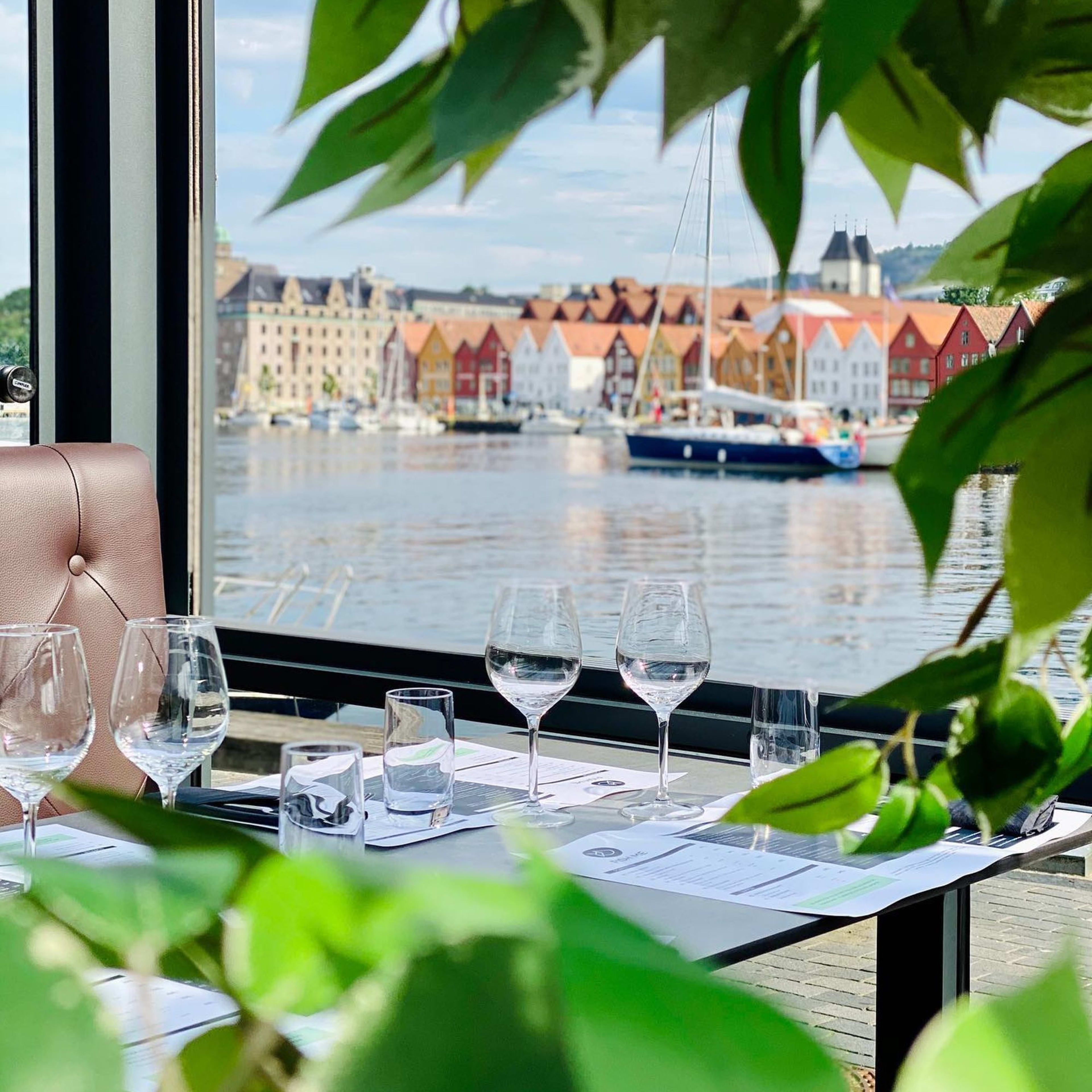 Ein Esstisch mit Weingläsern, Menüs und Servietten mit Blick auf das Wasser in Bergen, mit historischen Gebäuden.