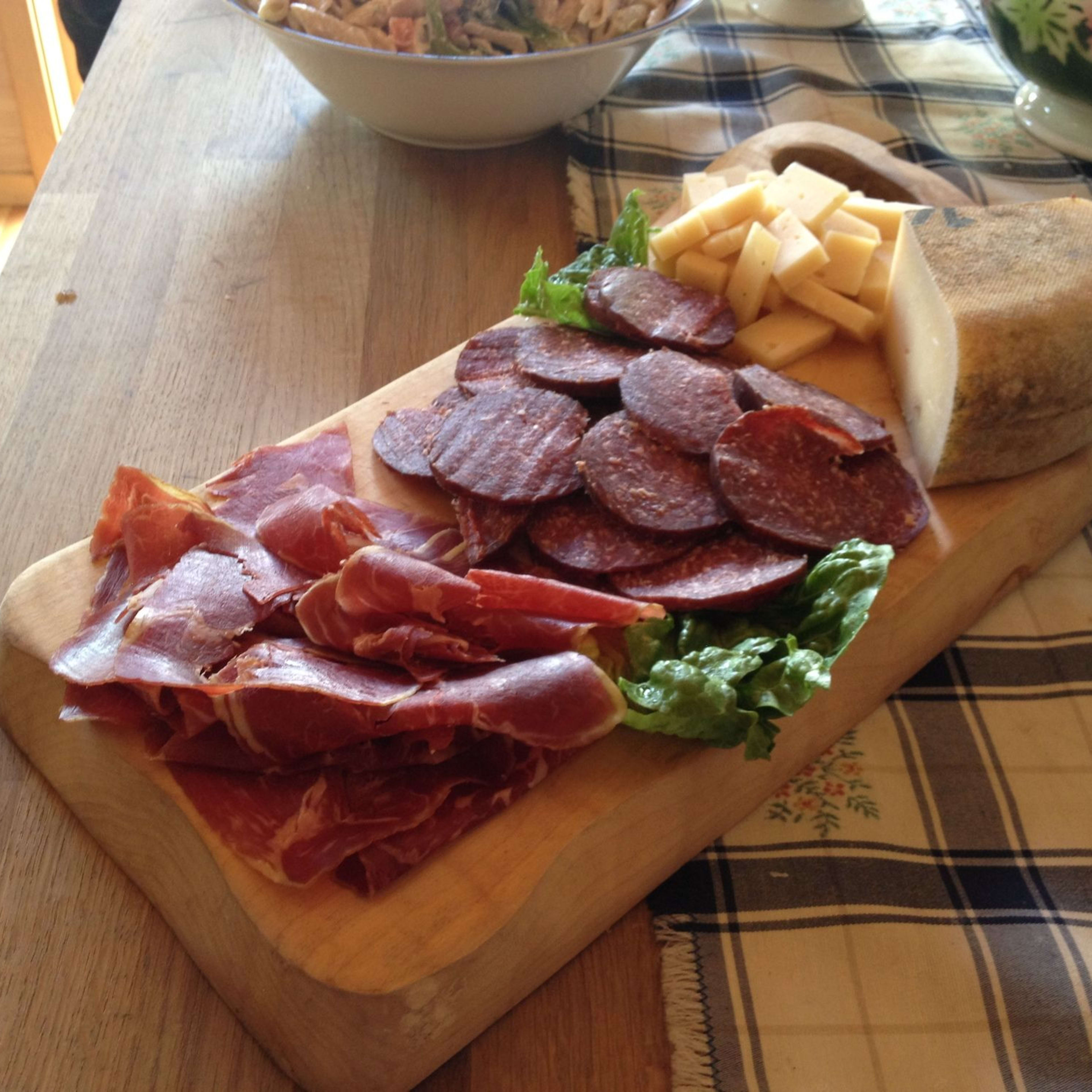 Rustikales Holzbrett mit Prosciutto, Salami, Käse und Salat auf einem karierten Tuch mit Salatschüssel im Hintergrund.