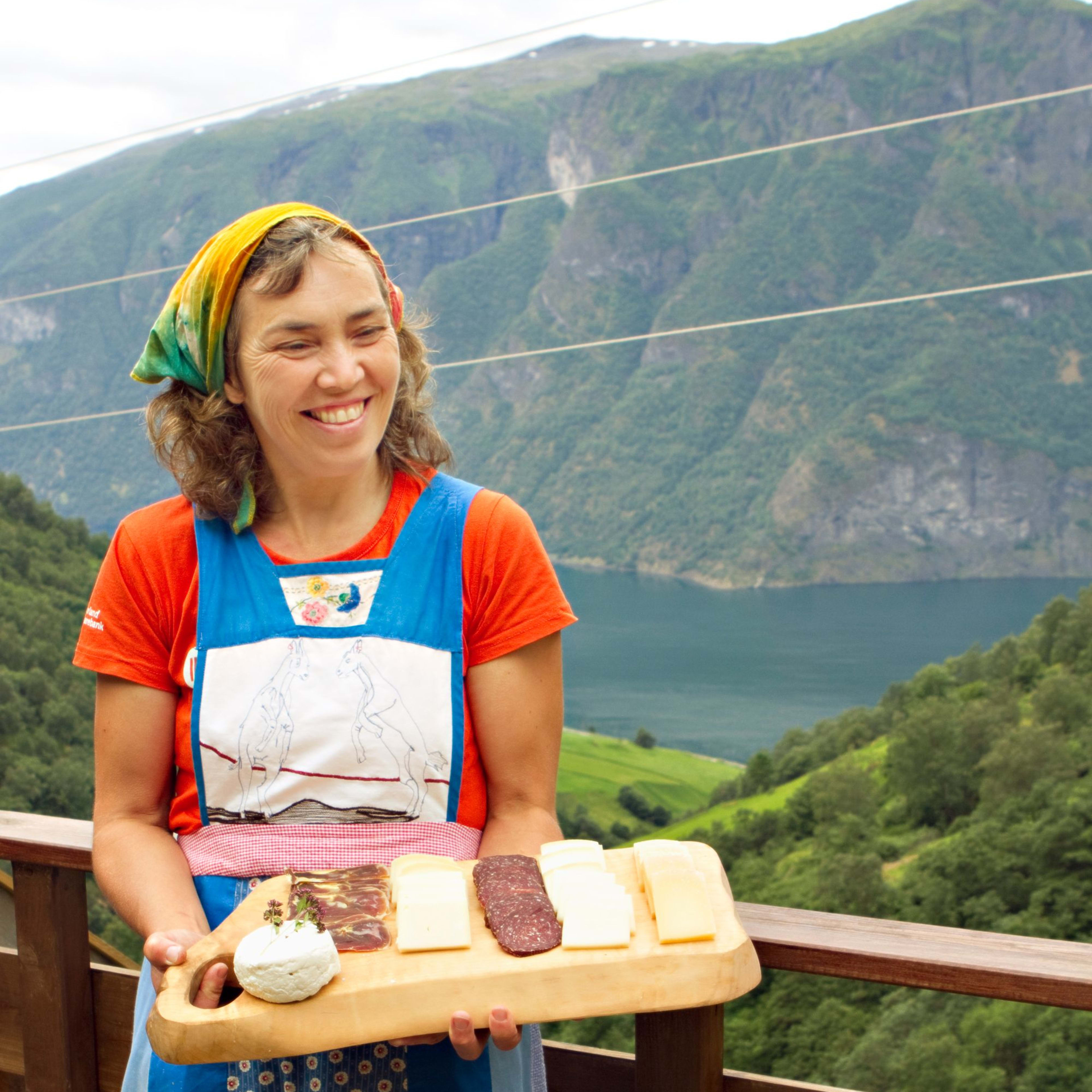 Lächelnde Frau hält ein Brett mit Käse, Schinken und frischem Käsebällchen, mit Blick auf Fjordlandschaft.
