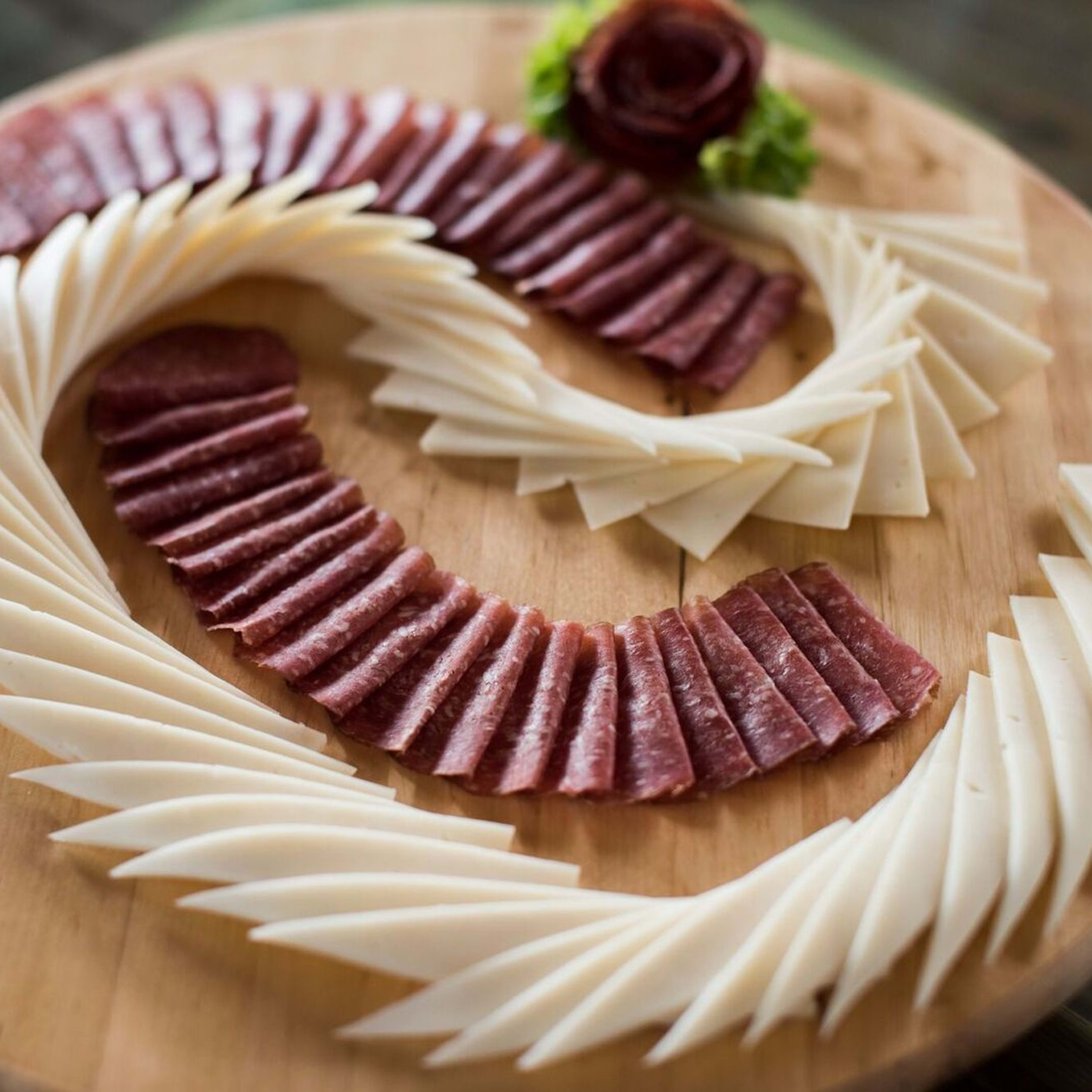 Charcuteriefat arrangert med spekemat, oster, salat og dekorativt rosenformet kjøttsentrum på trebrett.