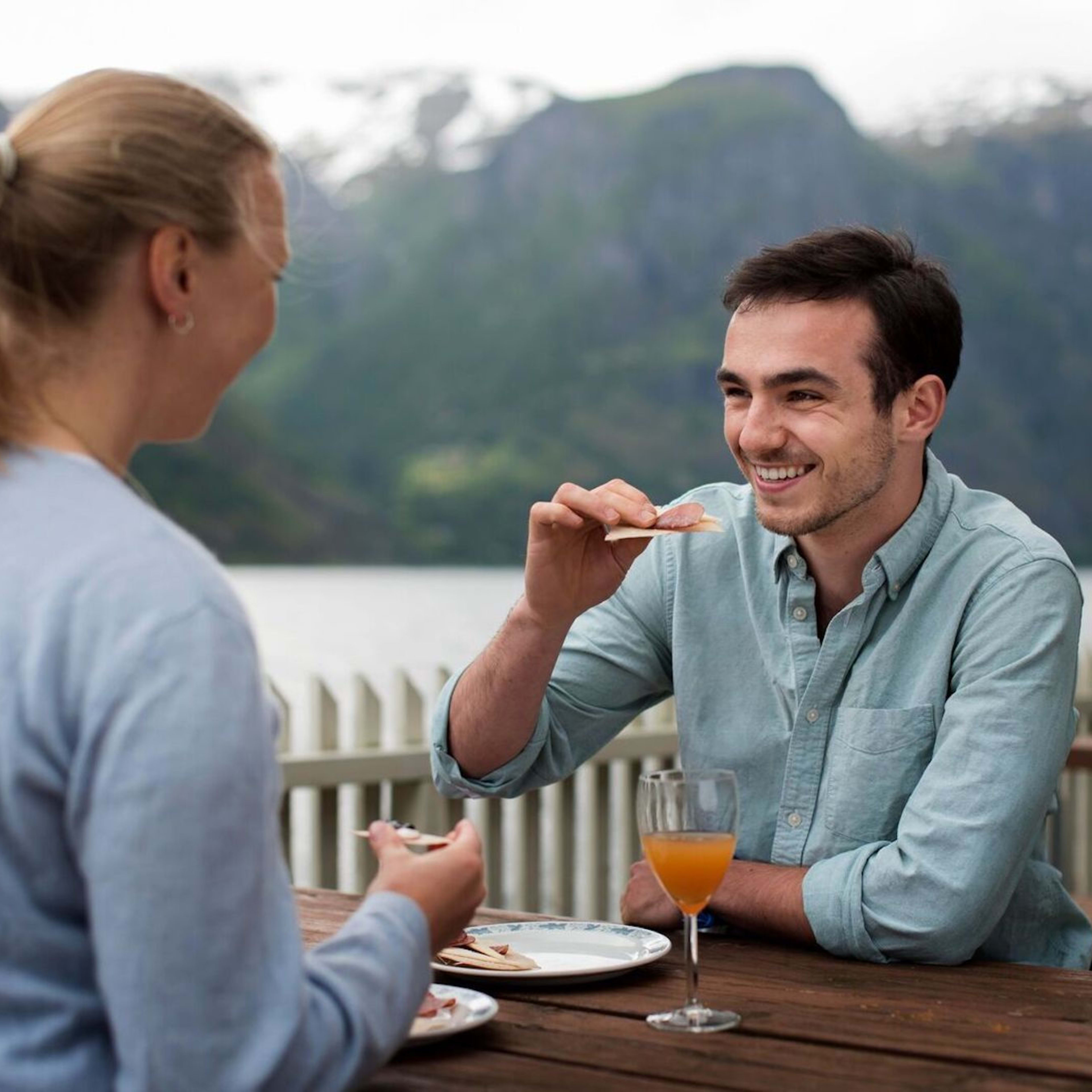 To personer som nyter et avslappet måltid utendørs ved fjorden, med lokal ost, spekemat og drikkevarer.