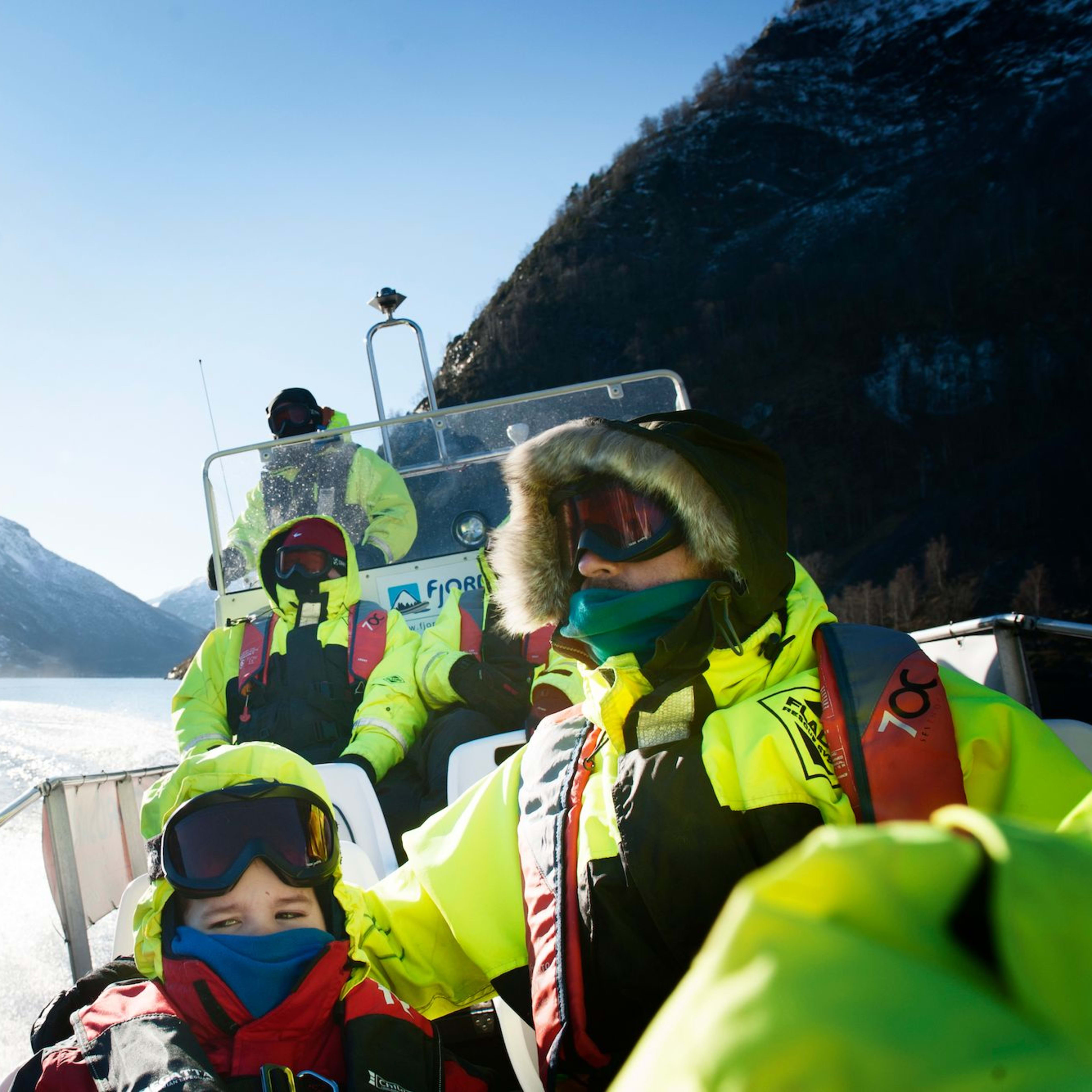 Menschen in Kälteschutzkleidung auf einem Boot durch einen sonnigen Fjord, umgeben von schneebedeckten Bergen und glitzerndem Wasser.