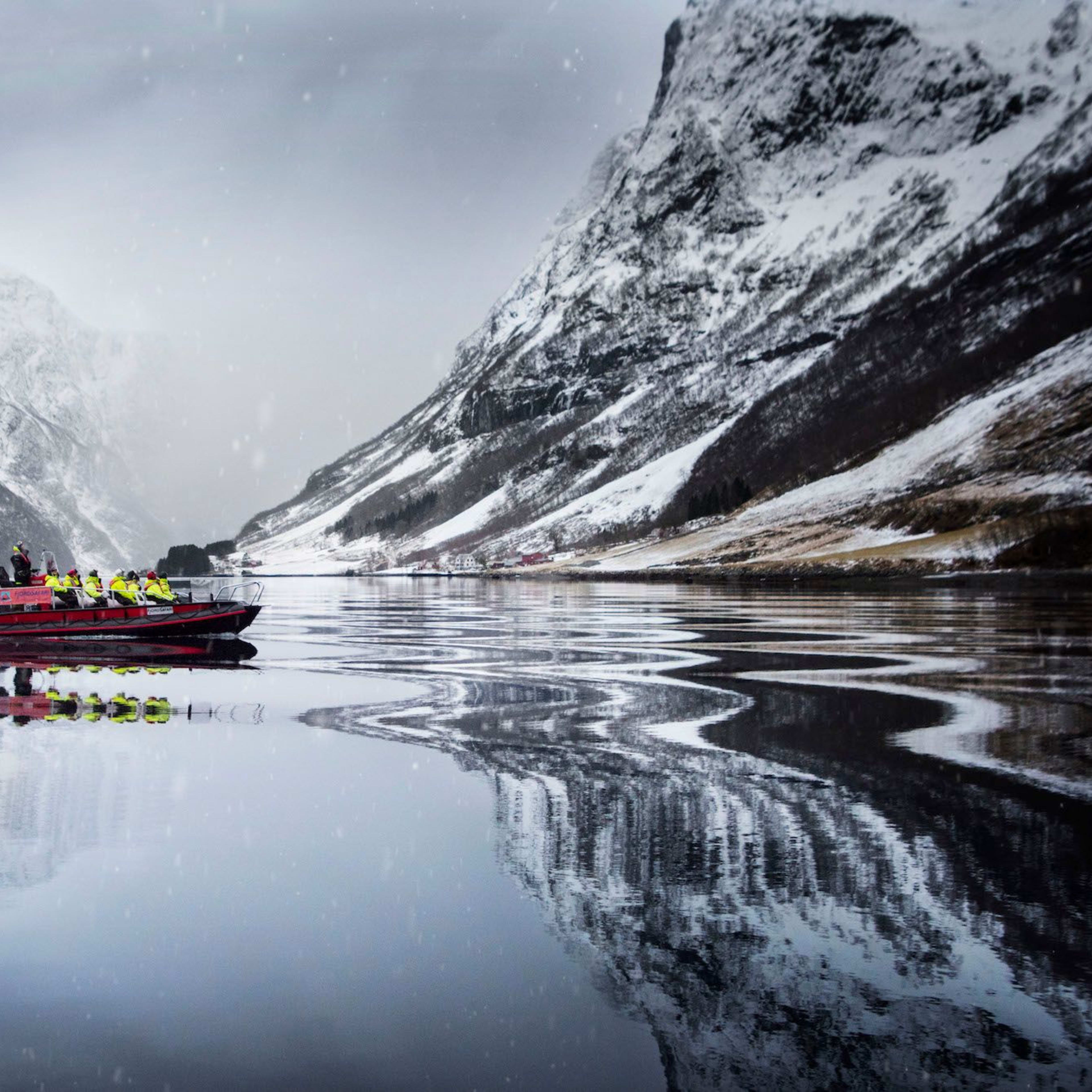 Rotes Schnellboot mit Passagieren in gelben Jacken gleitet über spiegelglattes Fjord, umgeben von schneebedeckten Bergen.