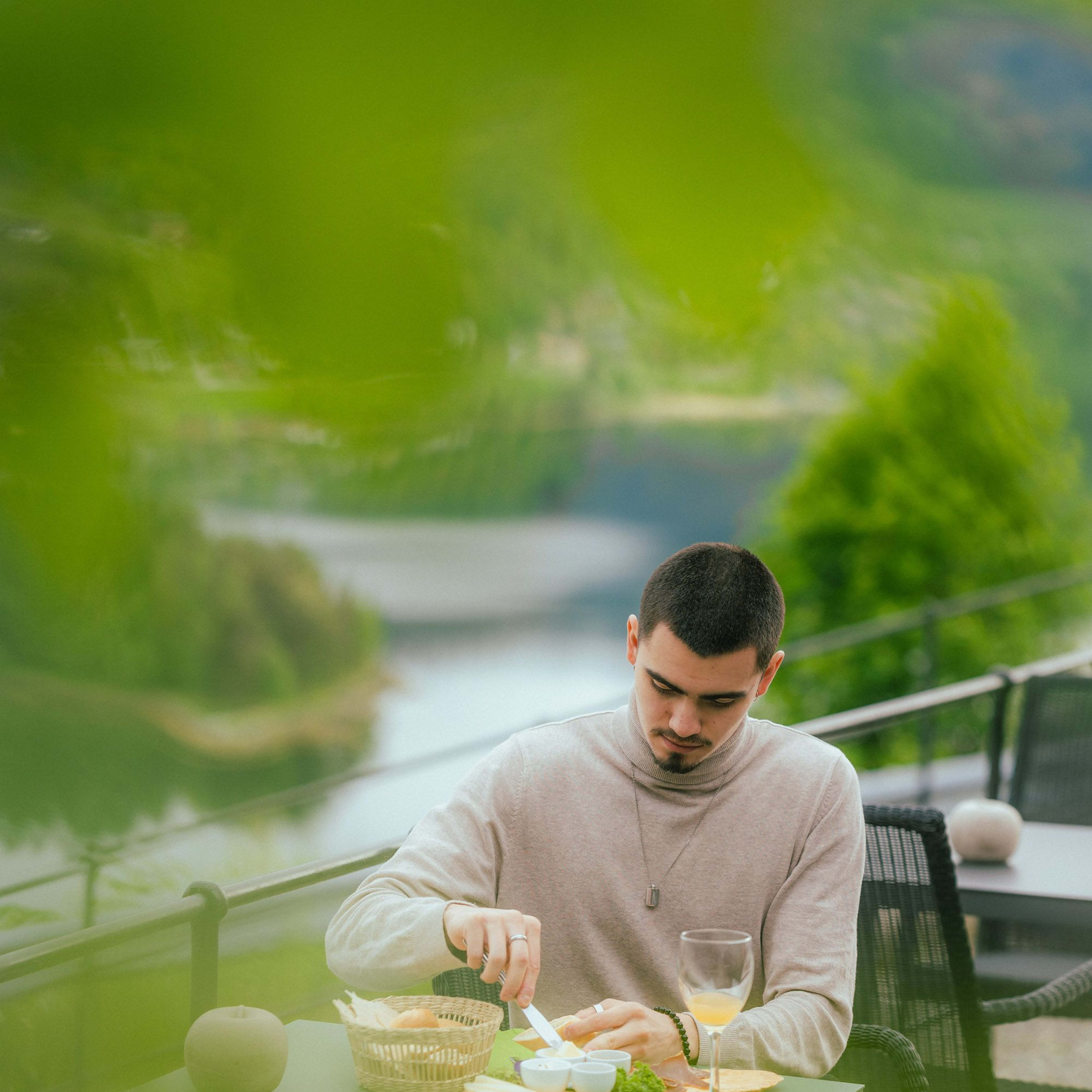 Mann smører smør på brød fra trebrett utendørs, med cider, sjøutsikt og grønn bakgrunn.