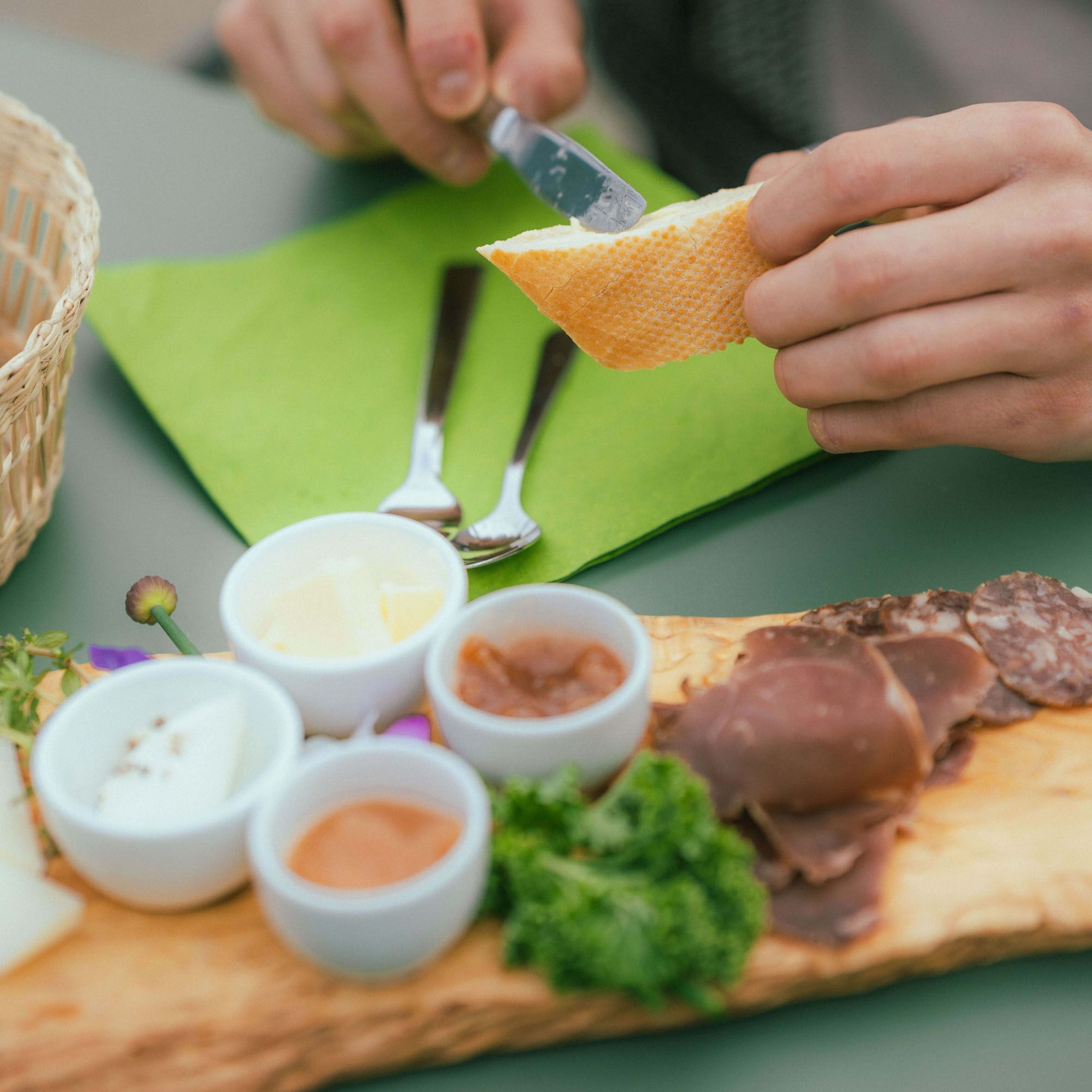 Person fokusert på å smøre smør på brød fra trebrett, med små skåler og spekemat i rustikk setting.