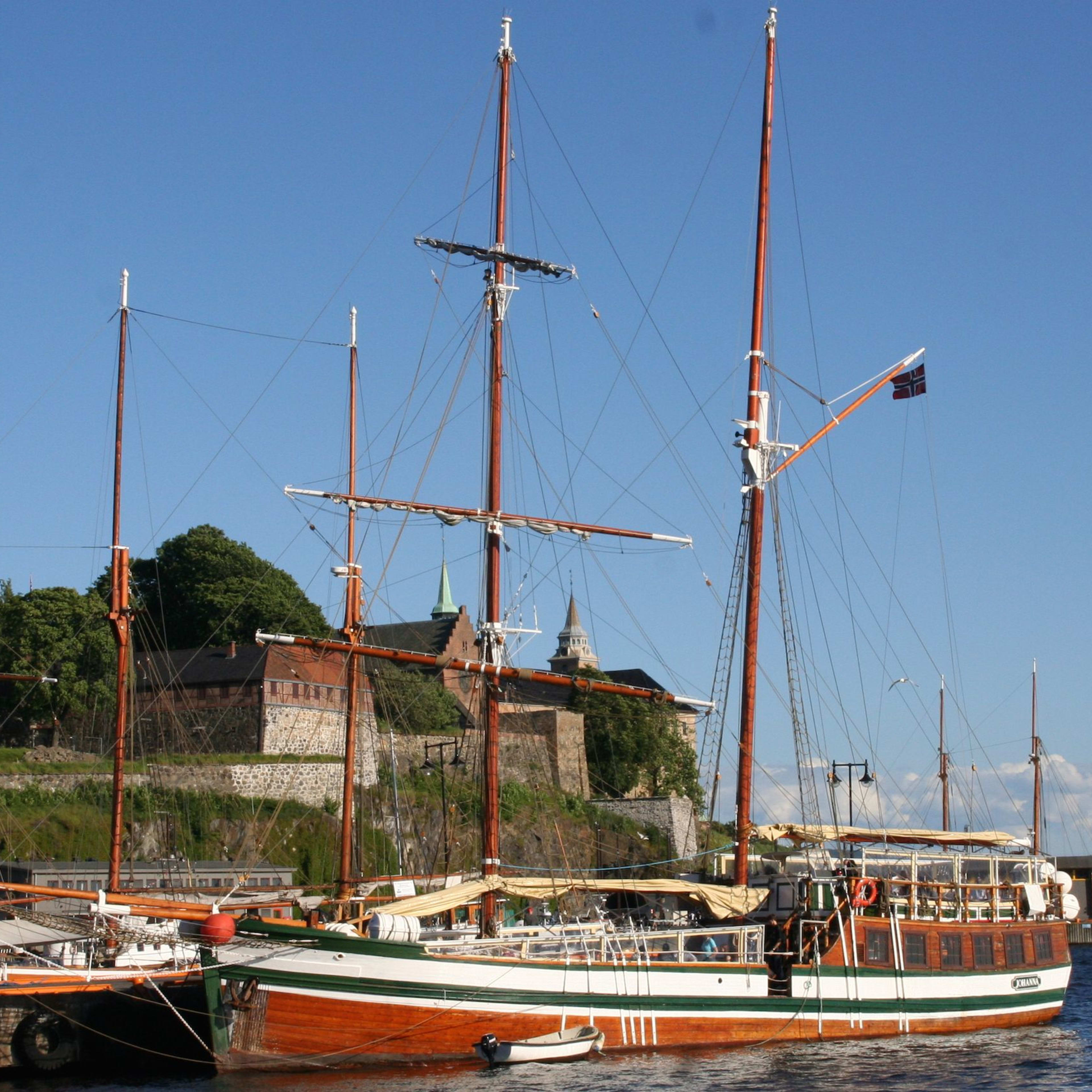 Wooden sailboat with polished masts docked near a historic fortress and green hills, with seagulls flying above.
