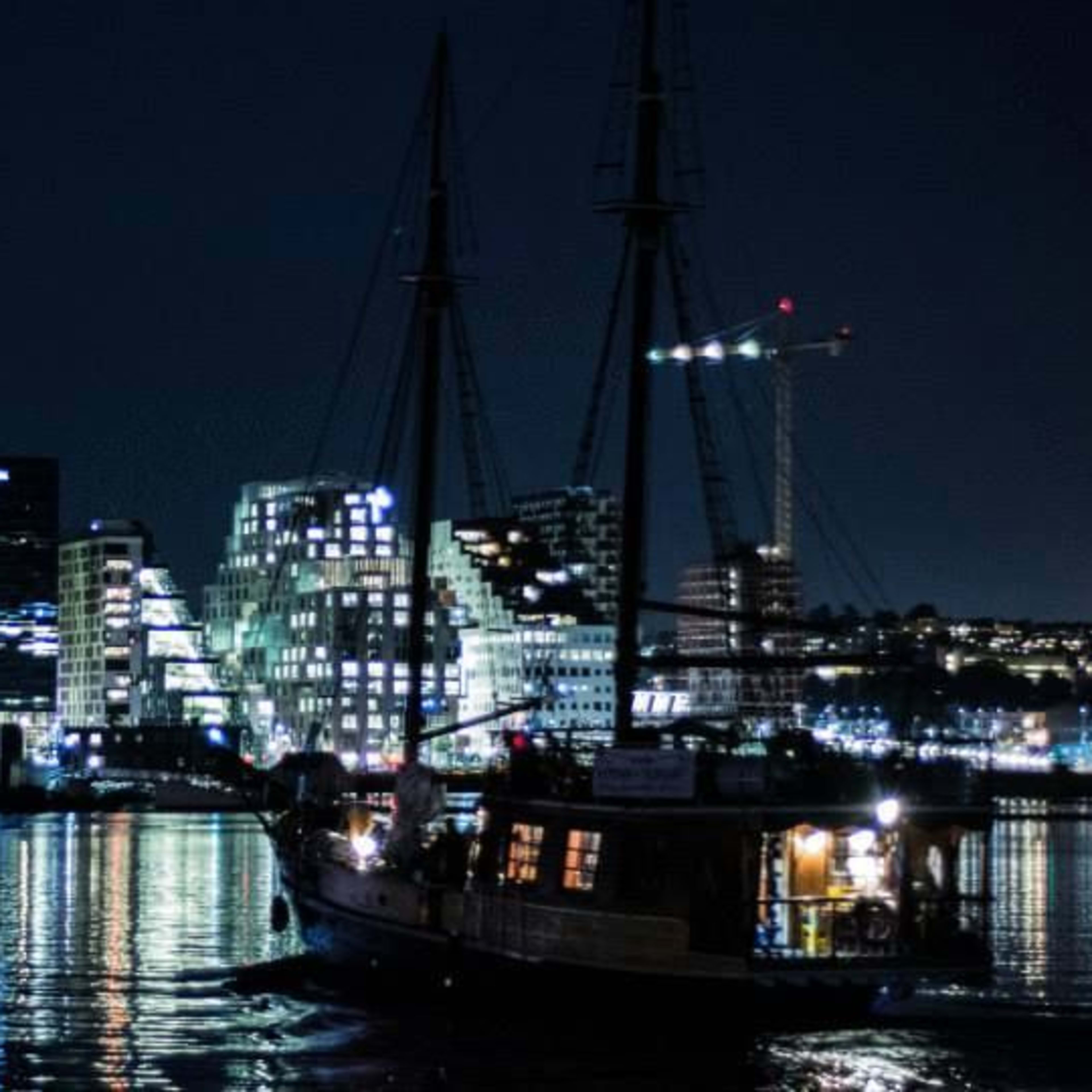 Nighttime cityscape with illuminated high-rise buildings and a sailboat in the foreground against a dark sky.