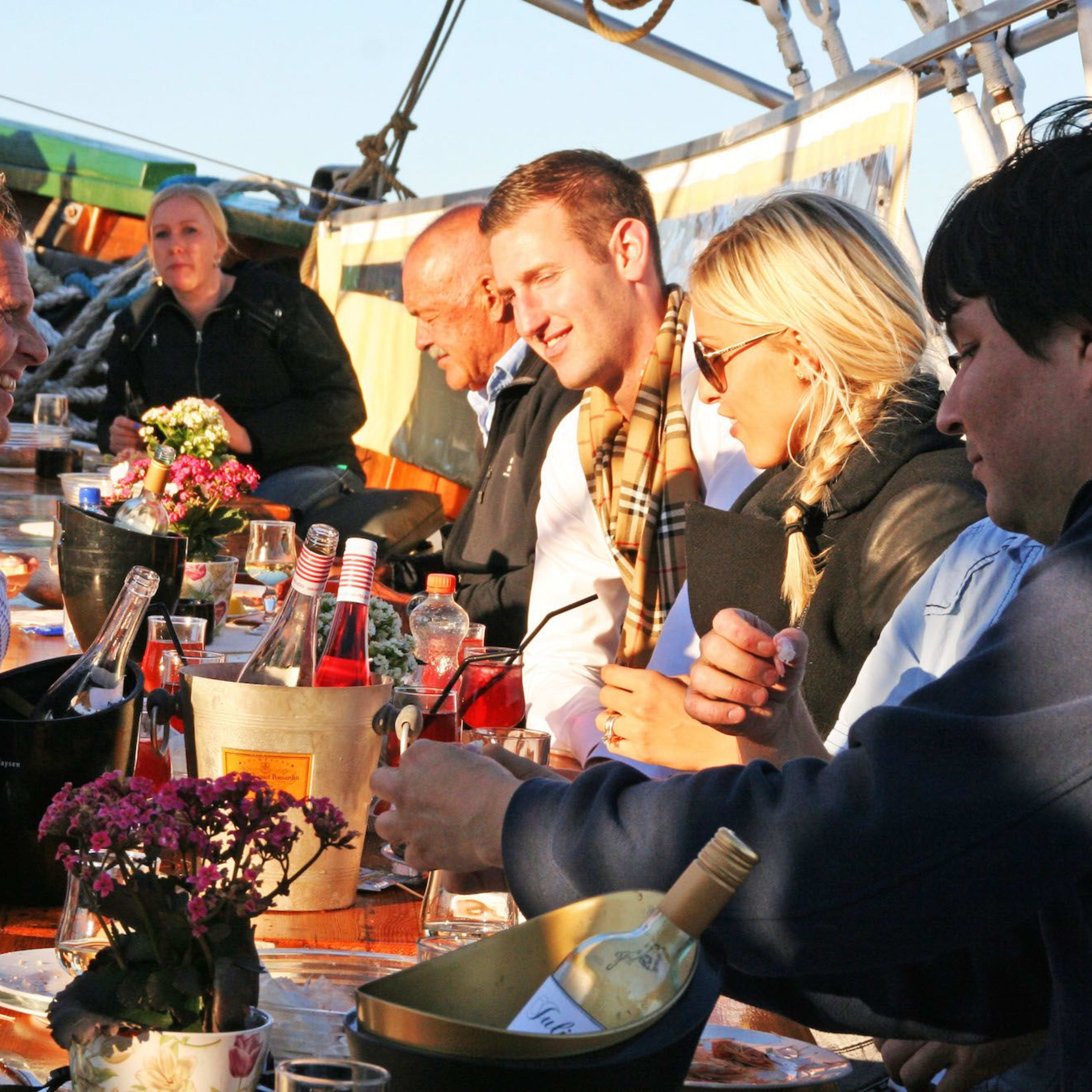 Group of people enjoying drinks on a boat deck, sharing smiles, with wine bottles and glasses, in warm sunlight.
