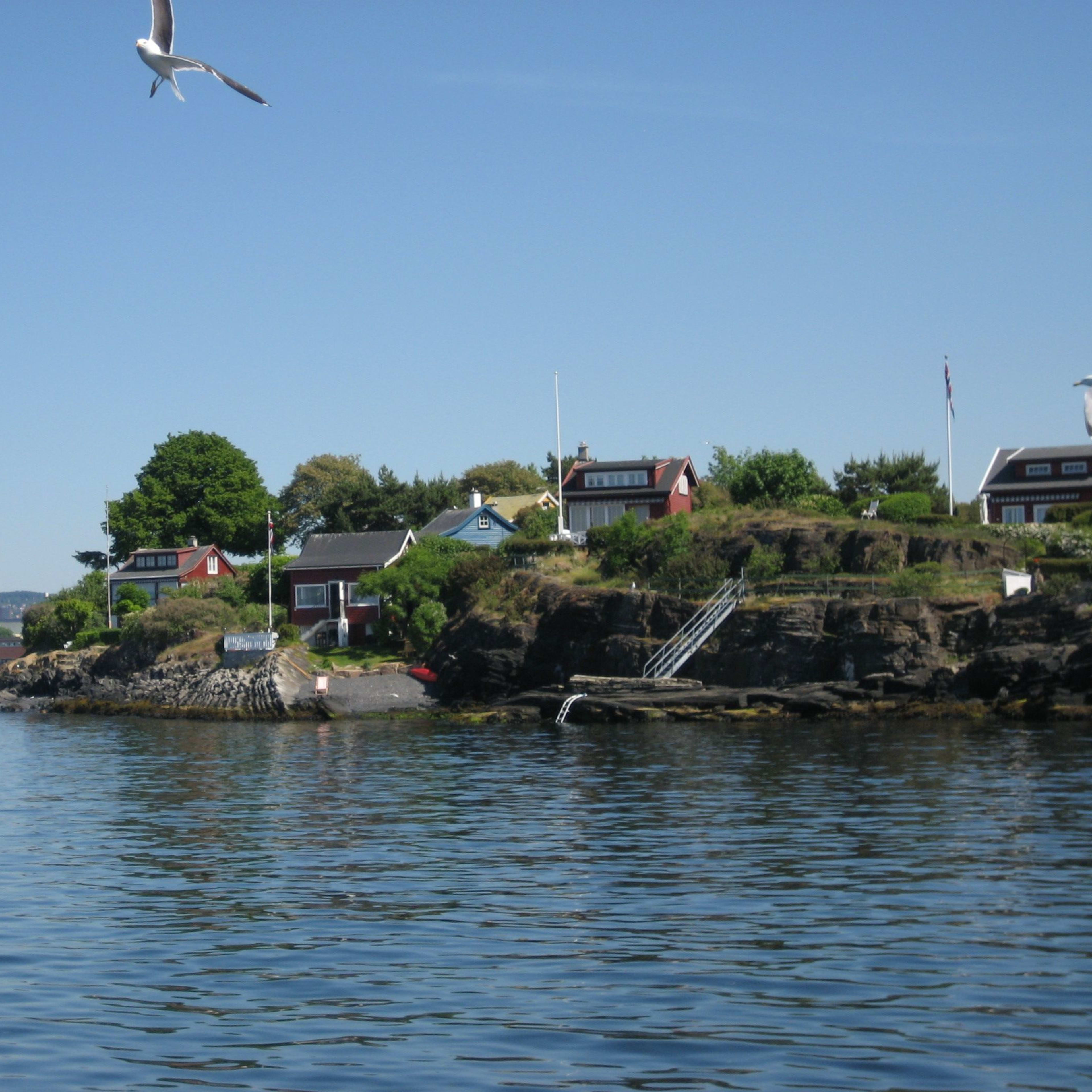 Red and white cottages on rocky cliffs with green trees, stairs to calm water, seagulls, and a small harbor.