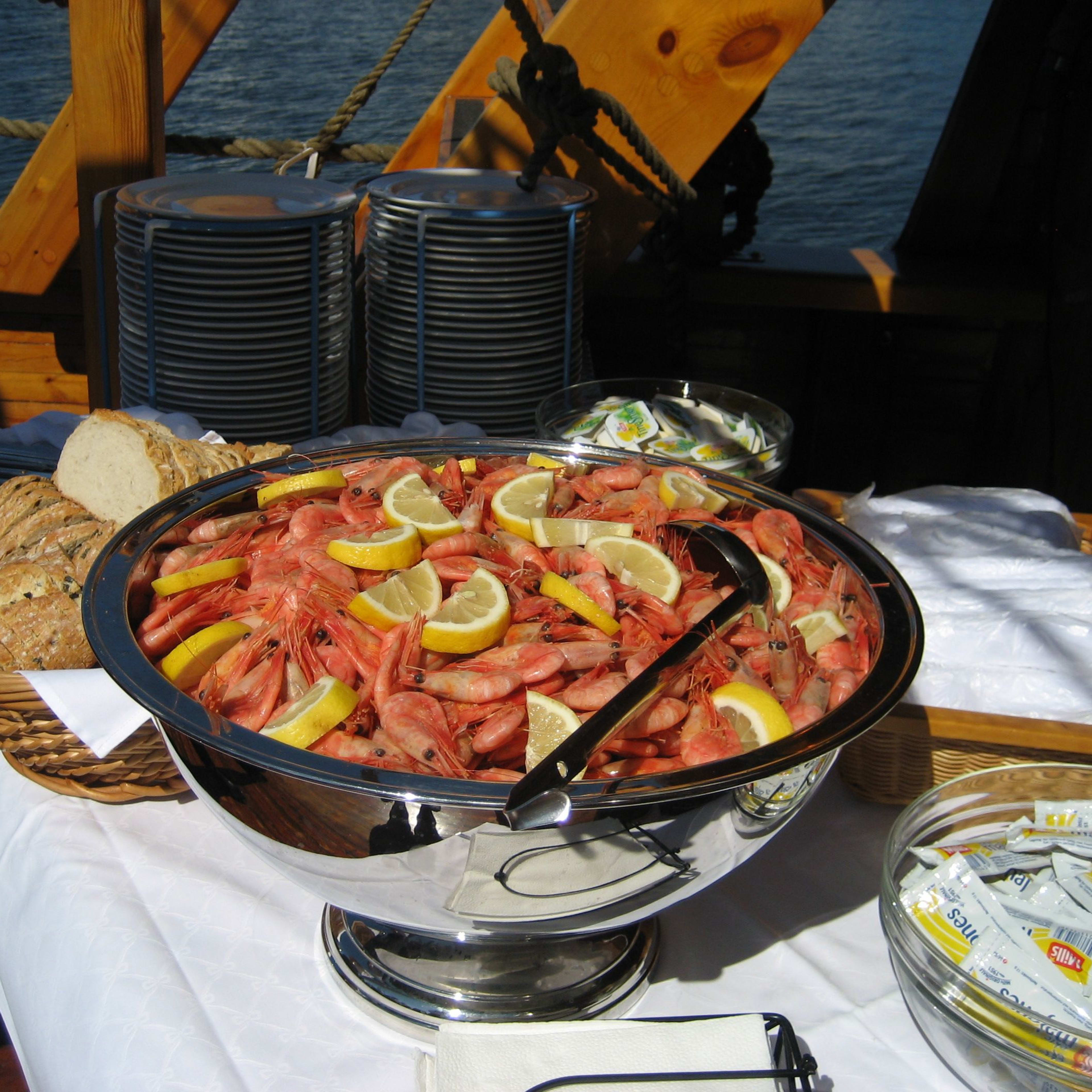 Silver bowl of shrimp with lemon wedges, surrounded by bread, plates, condiments, and napkins on a boat deck.