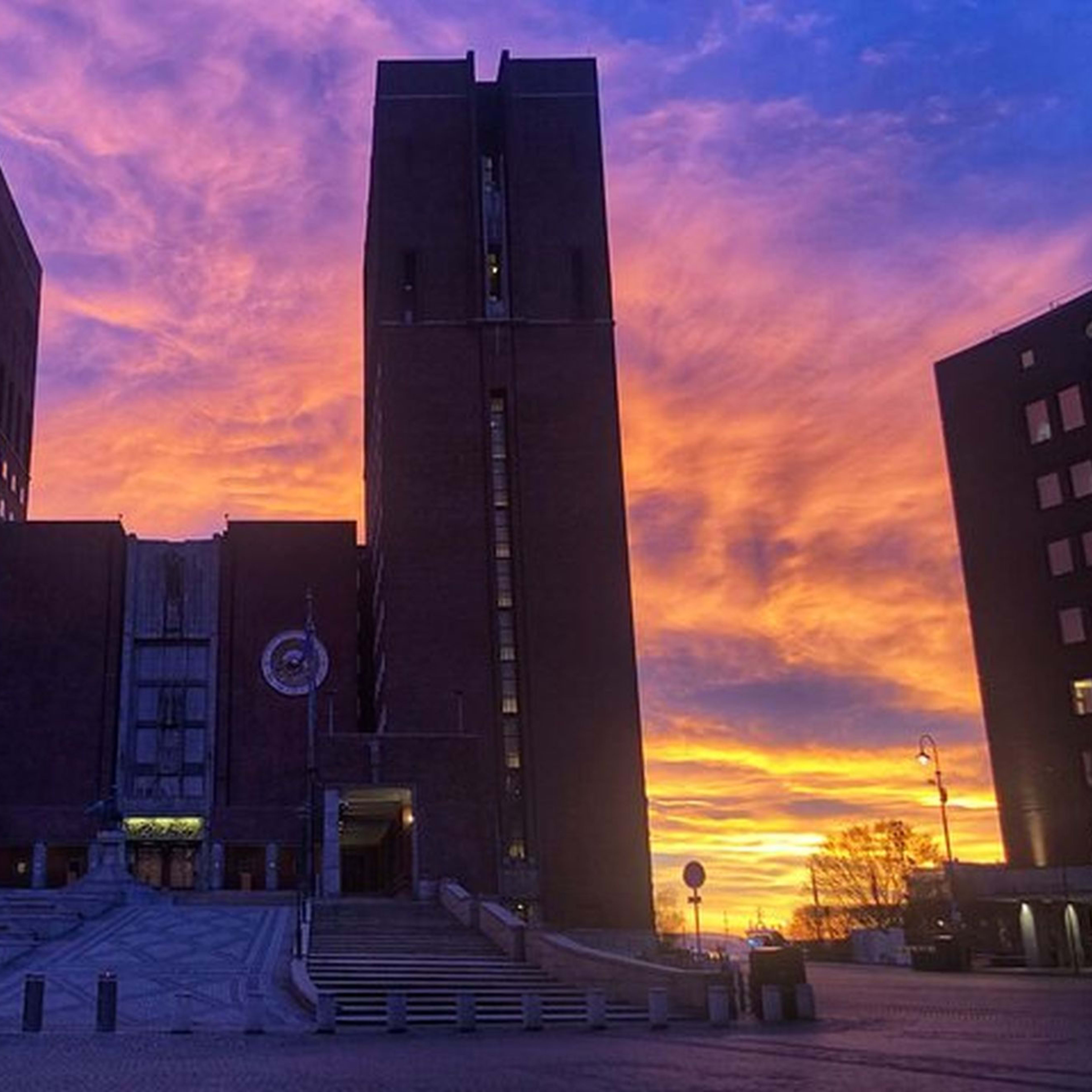 Dramatisk solnedgang med livlig lilla, rosa og oransje himmel over en symmetrisk murbygning, med et pyntet juletre.