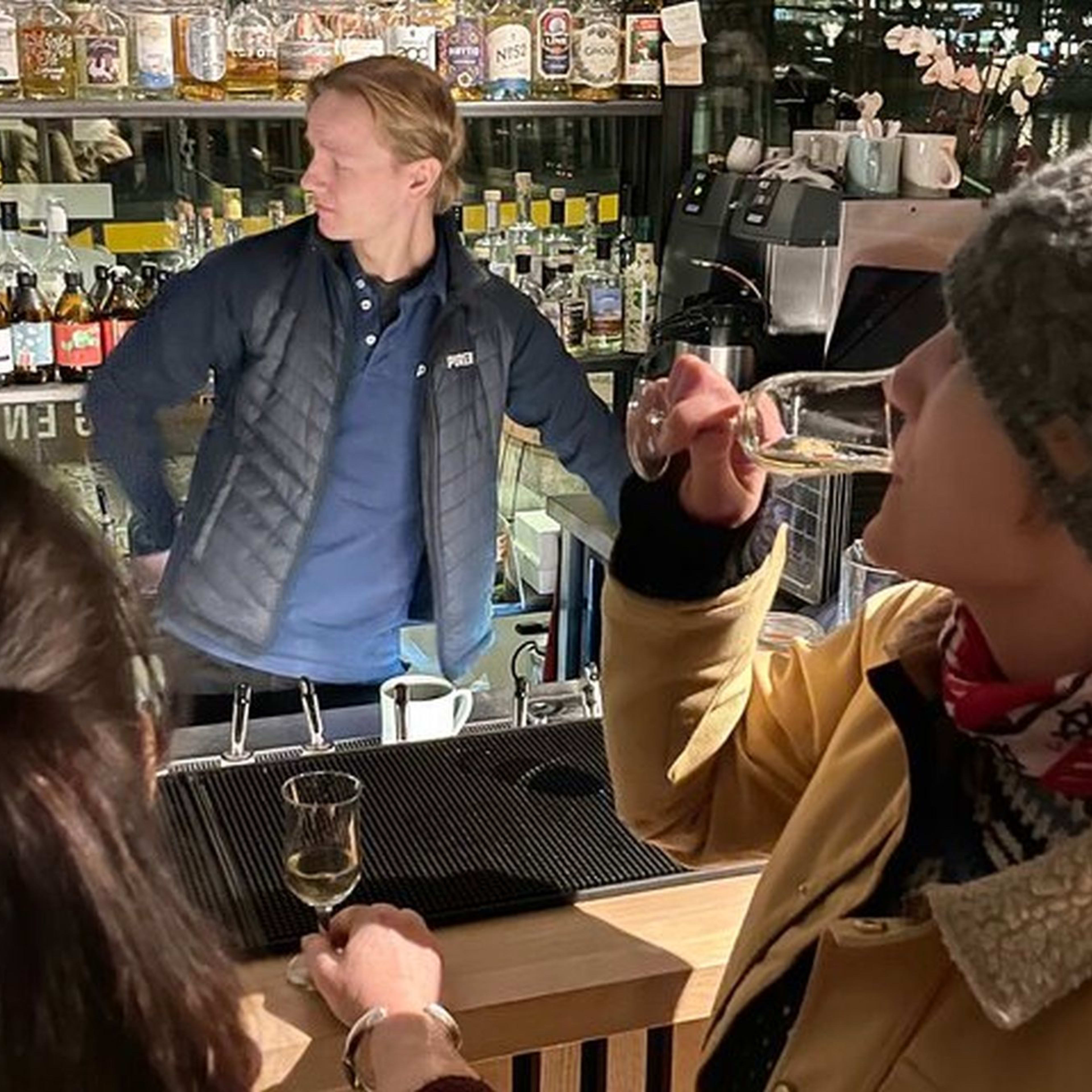 To kvinner på stilfull bar, den ene sipper på drinken mens den andre ser på, med bartender og hyller med flasker i koselig atmosfære.