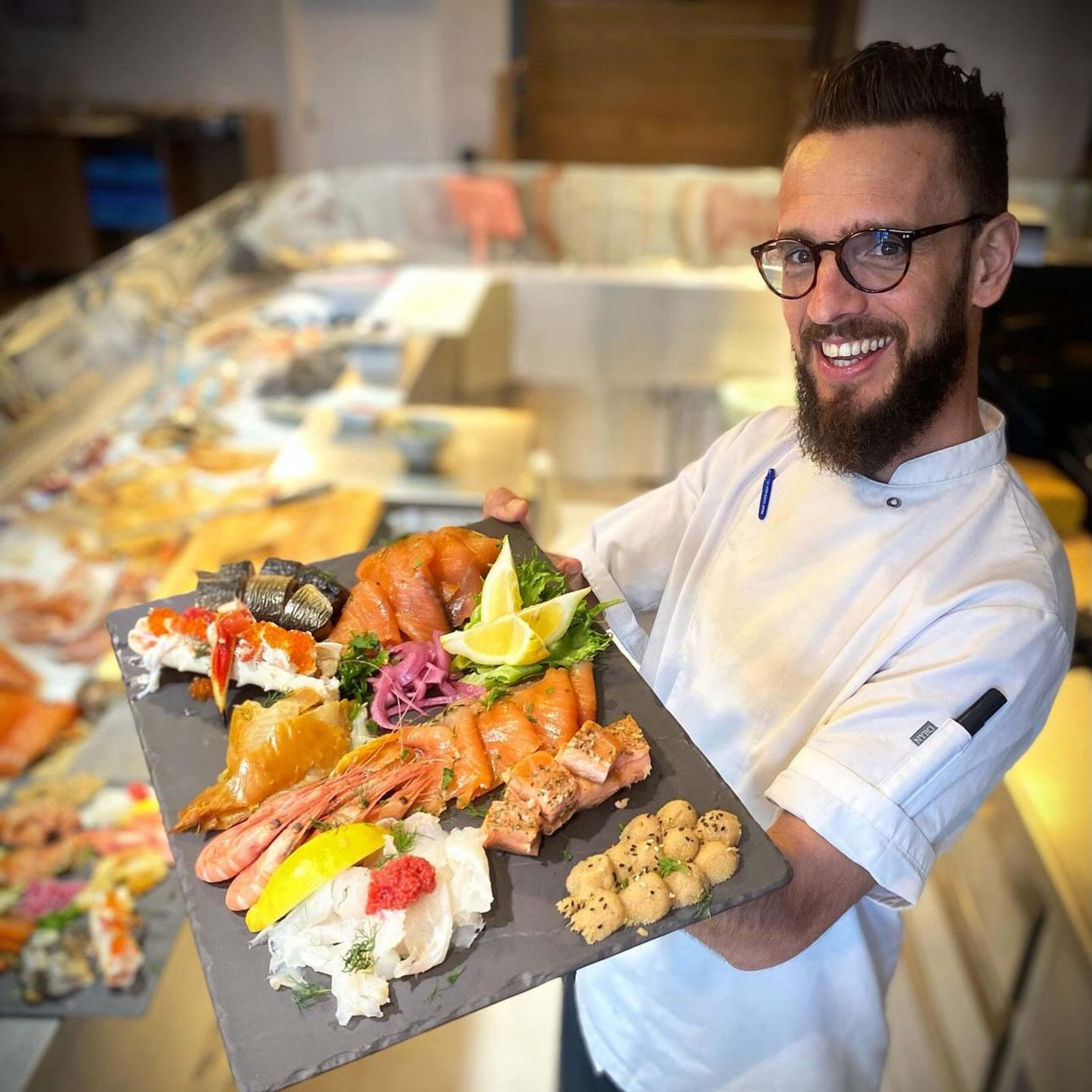 En smilende kokk som presenterer et fat med fersk sjømat, inkludert laks, reker, sushi, rogn, marinert sjømat og garnityr som sitronbåter og friske urter