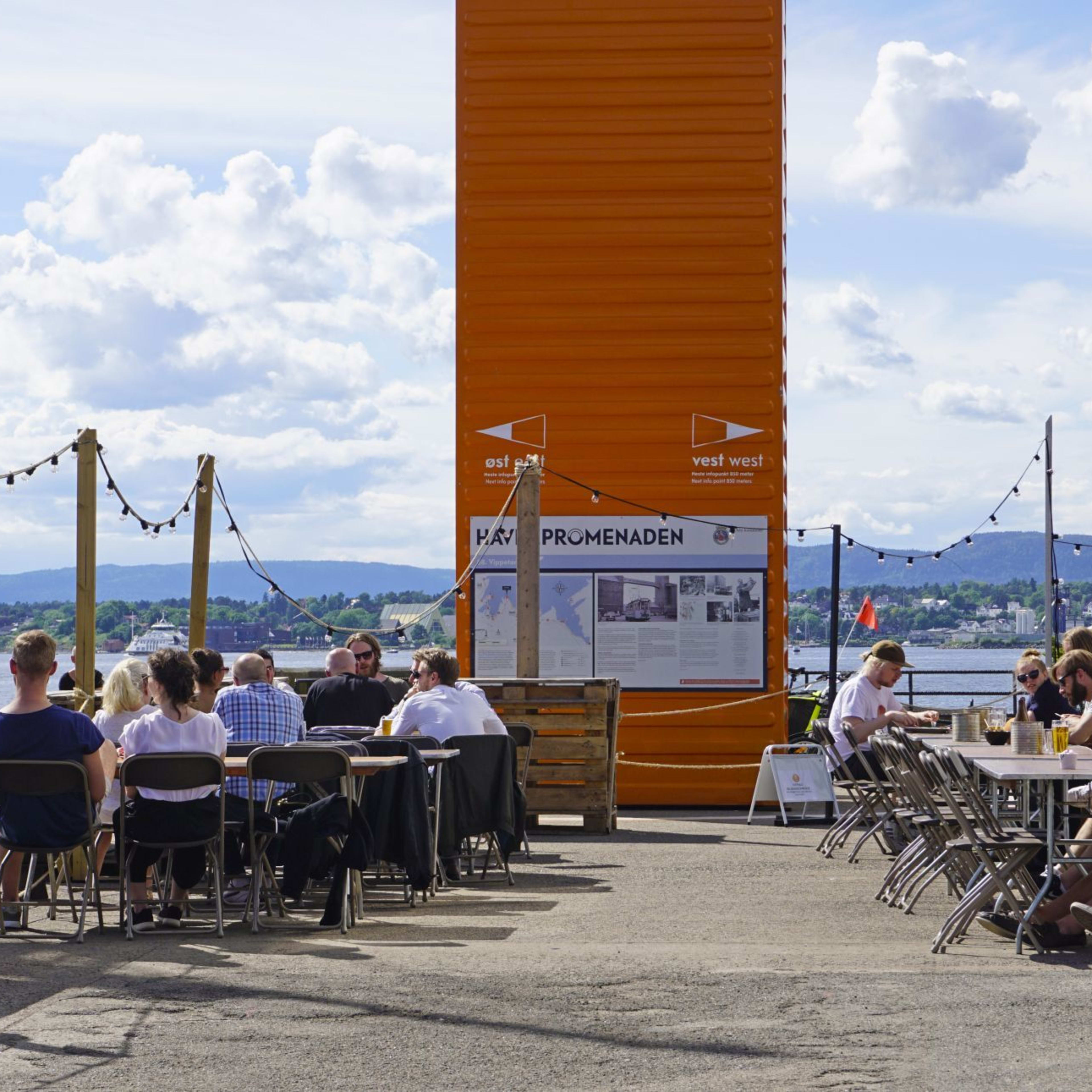 Et utendørs sitteområde ved vannet med rustikk tremøbler, lysslynger og en slående oransje fraktcontainer.