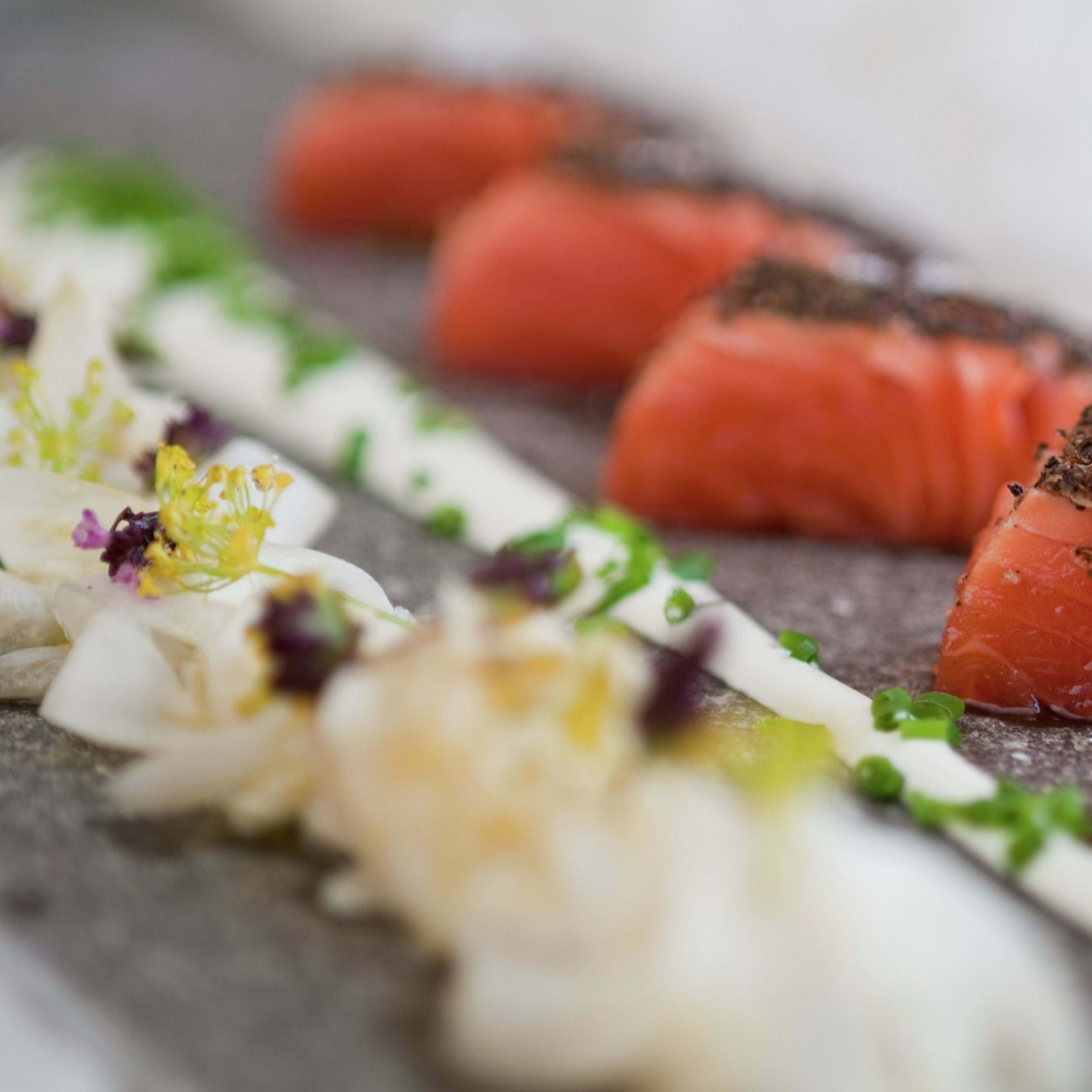 An elegant presentation of cured salmon with pepper crust, creamy sauce, and fresh herbs on a stone plate.