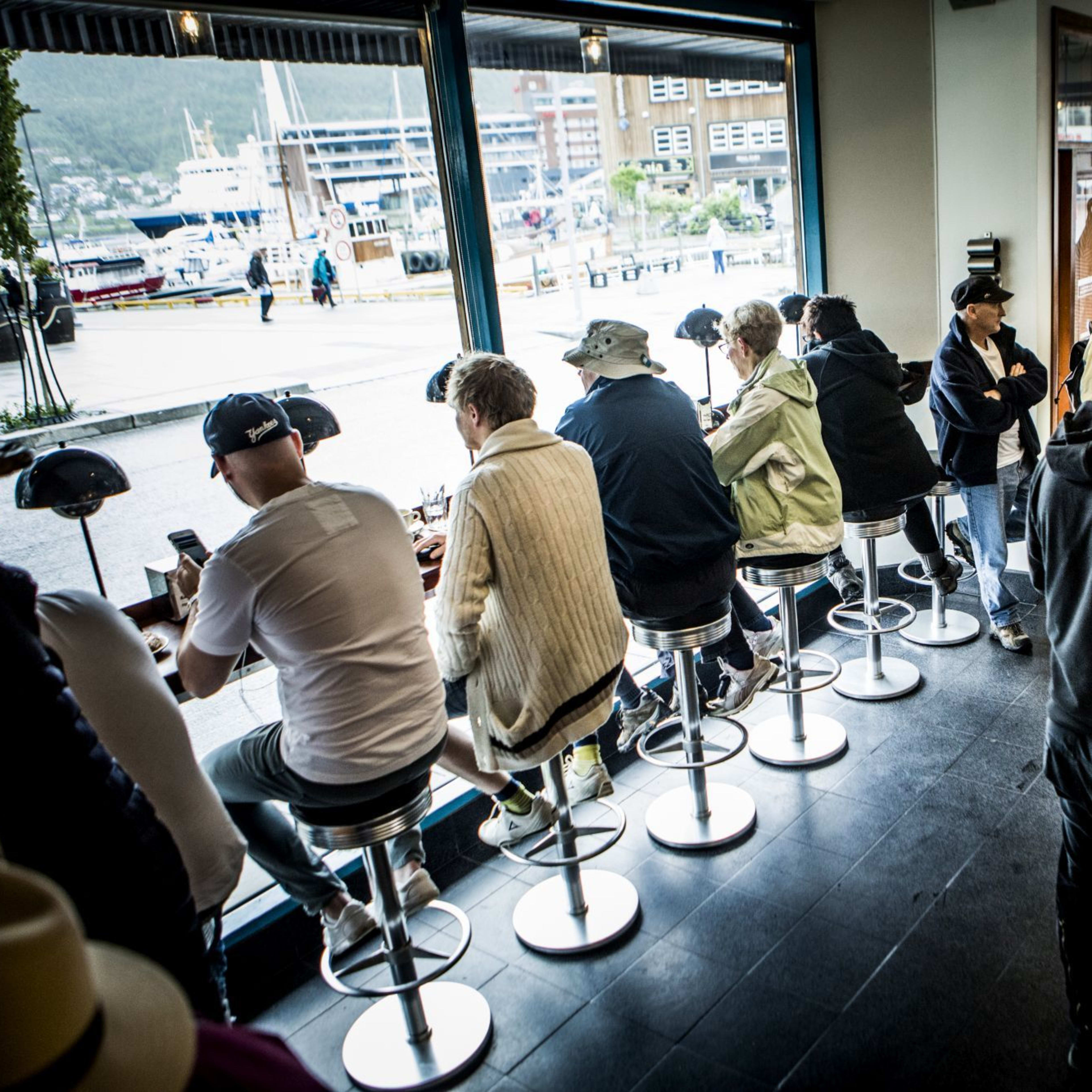 A bustling coffee shop with large windows, customers enjoying drinks, and a modern interior with metal stools and lighting.