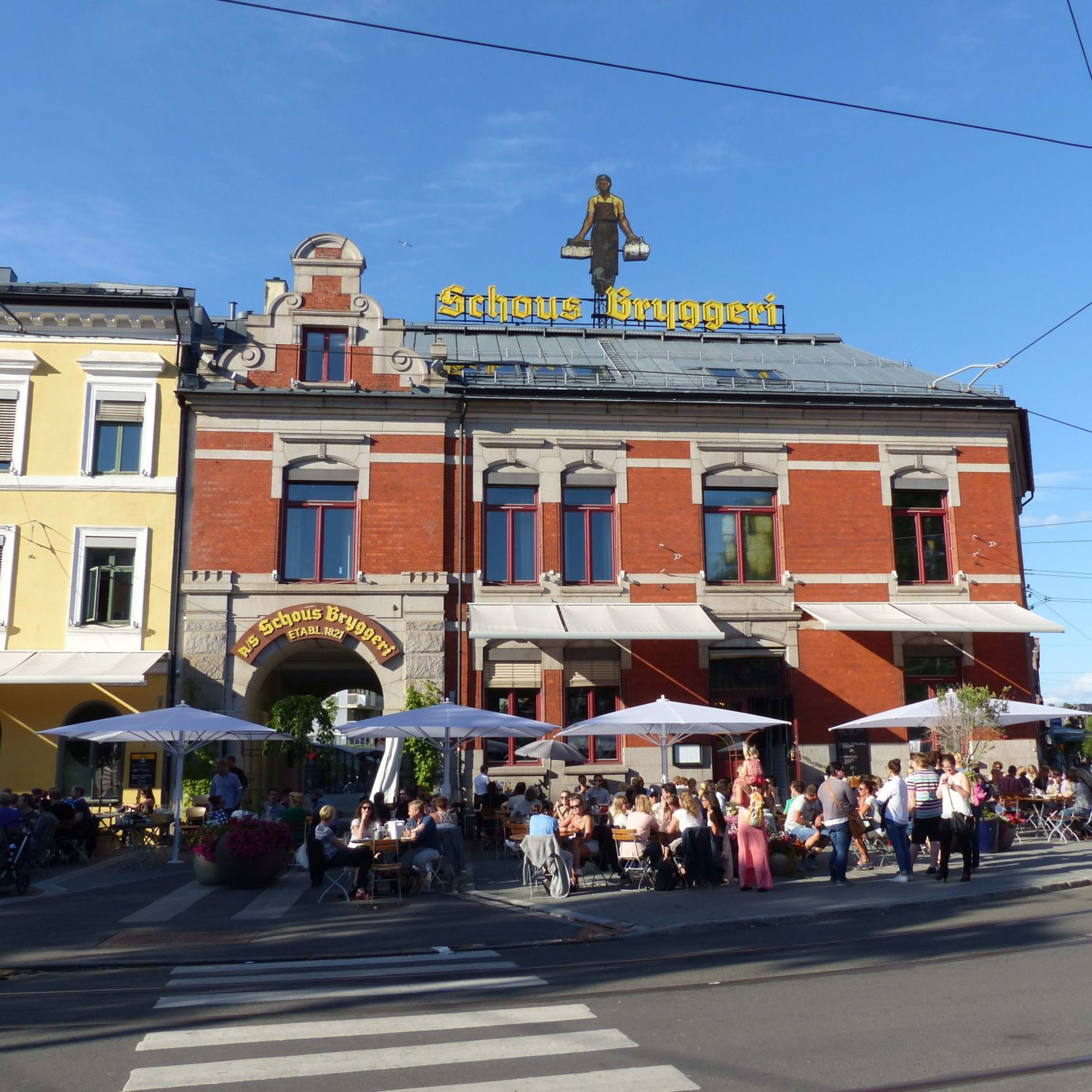 Lebendige Außenszene bei "Schous Bryggeri" mit Menschen, die Getränke unter Sonnenschirmen genießen, rotes Backsteingebäude und klarer Himmel.